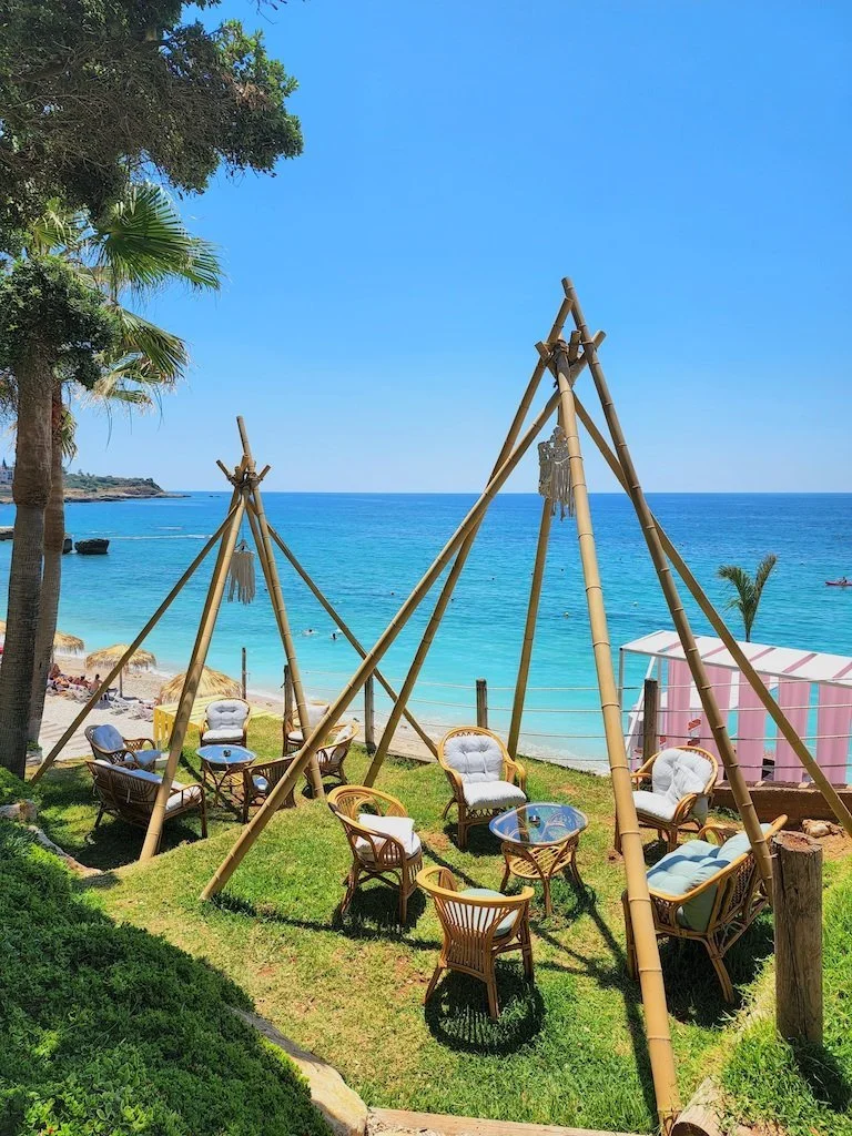Outdoor seating area with bamboo structures and cushioned chairs on grassy area by the beach, with the ocean and blue sky in the background.