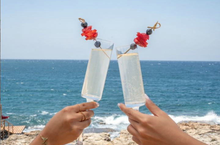 Two hands holding champagne flutes with skewered berry and candy garnishes, clinking together against a beach ocean background.