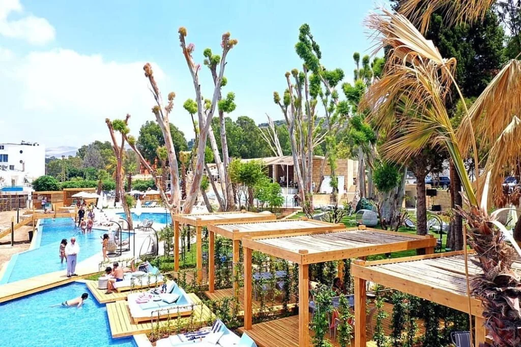 A lively outdoor pool scene with people relaxing and swimming, surrounded by wooden pergolas, tall trees, and tropical plants under a partly cloudy sky.