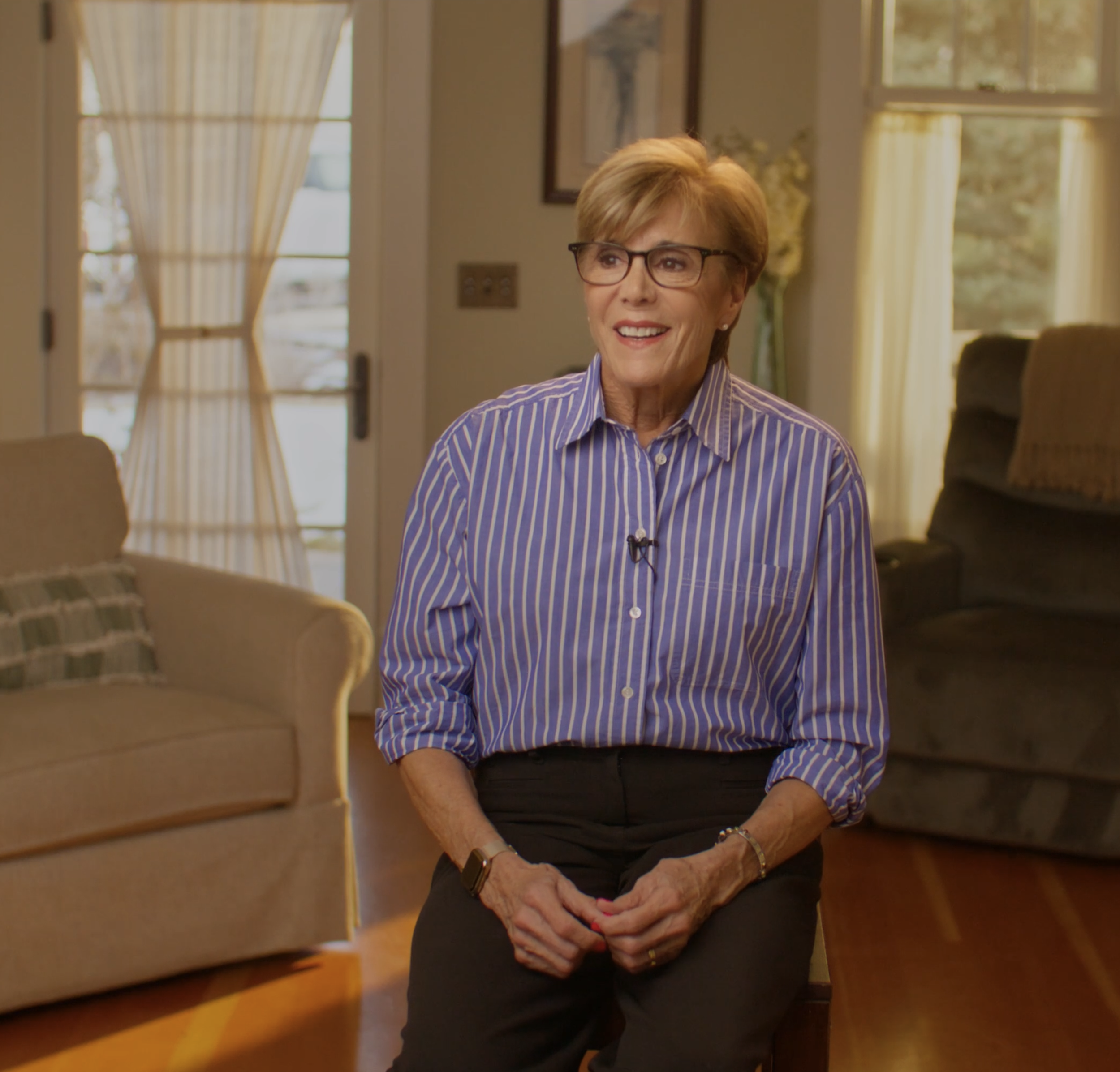 An elderly woman with short gray hair, glasses, and a striped button-up shirt, sitting indoors with large windows in the background. She is smiling and holding her hands over her abdomen.