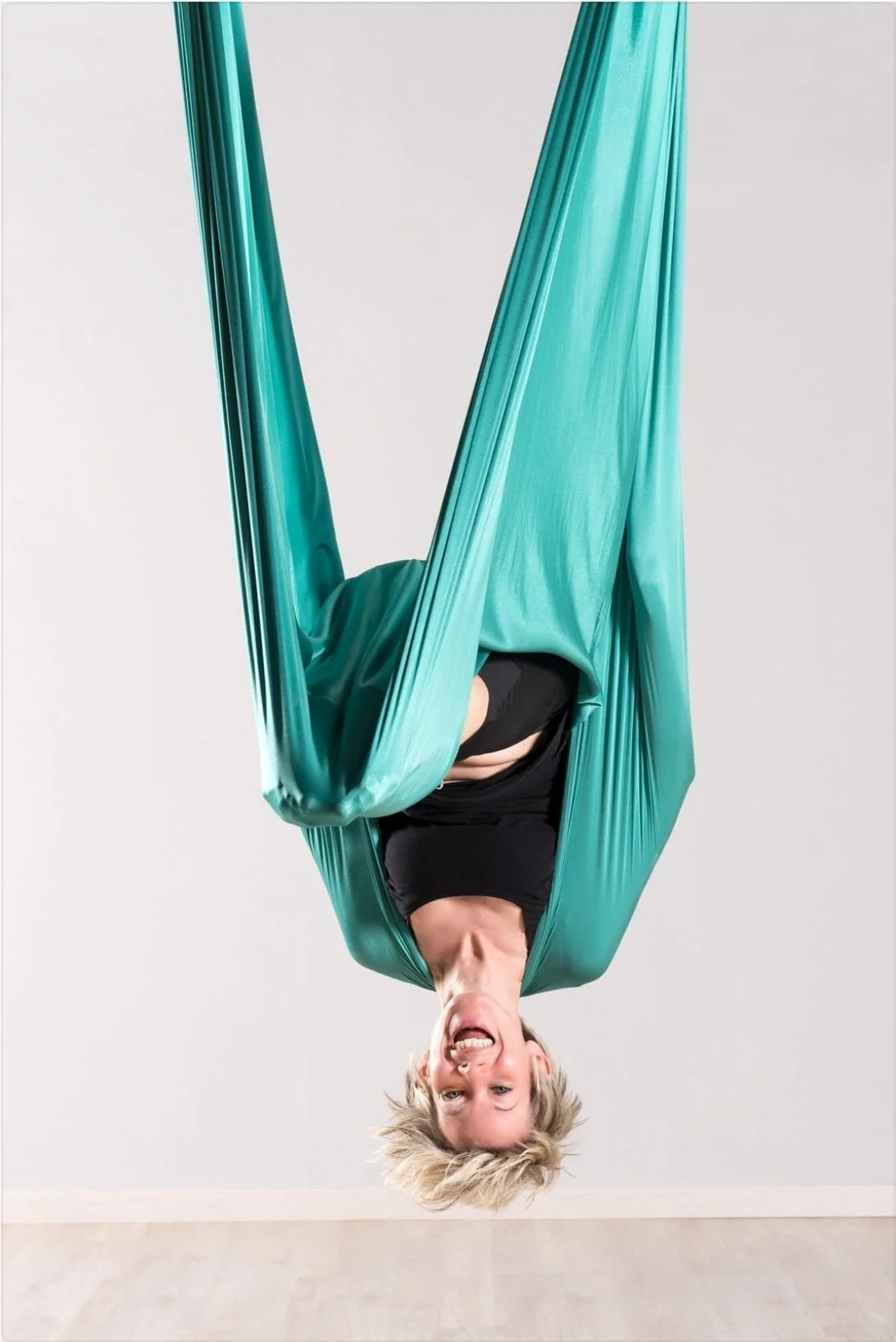 Person hanging upside down in a teal aerial silk hammock, smiling and making a silly face in a studio with a plain background.