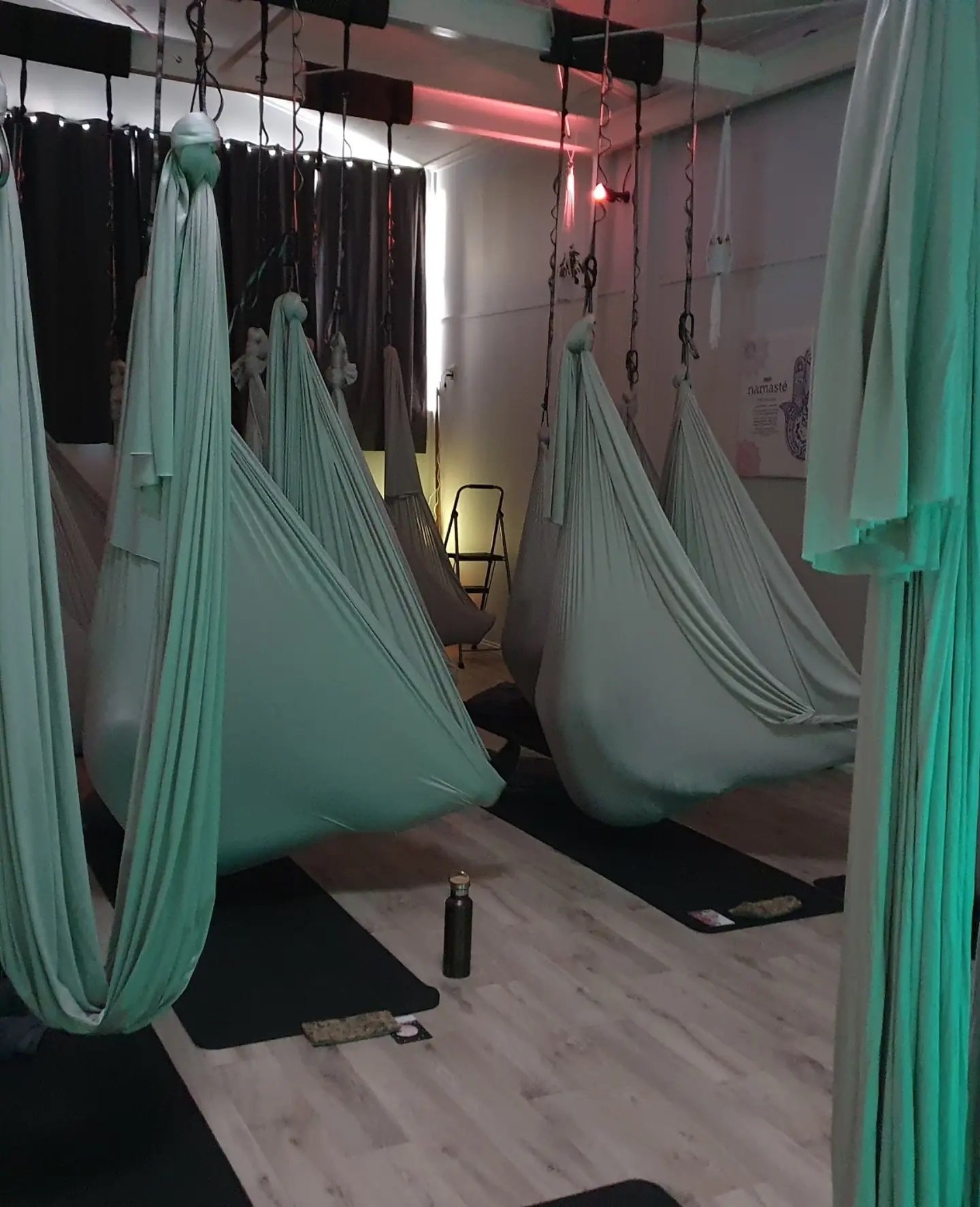 Indoor aerial yoga studio with hanging fabric hammocks, yoga mats, water bottles, and a glow light.