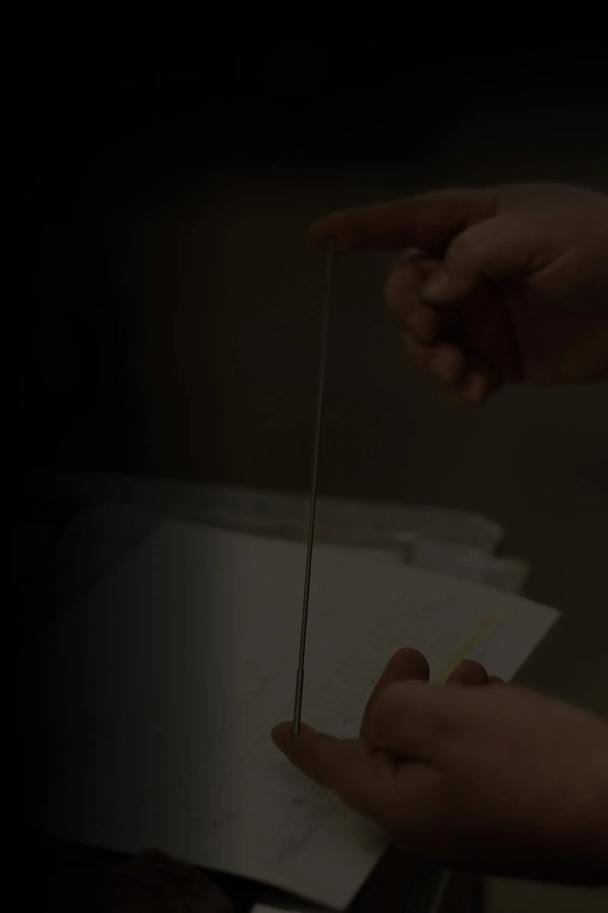 A person's hand holding a thin, straight metal rod or filament over a piece of paper or notebook.