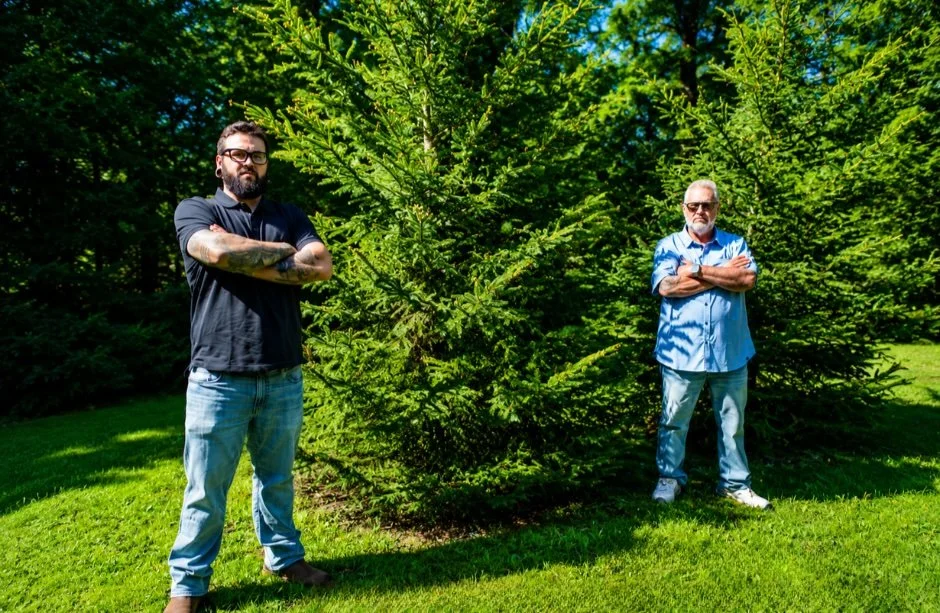 Mike and Mark Sampson, owners of Muskoka Pest Control, standing outdoors on a lawn surrounded by evergreen trees in Muskoka