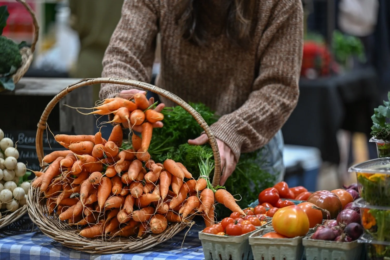 A person in a brown knit sweater arranging a wicker basket overflowing with fresh organic carrots at an indoor farmers market stall, surrounded by heirloom tomatoes, cherry tomatoes, and red onions on a blue and white checkered tablecloth