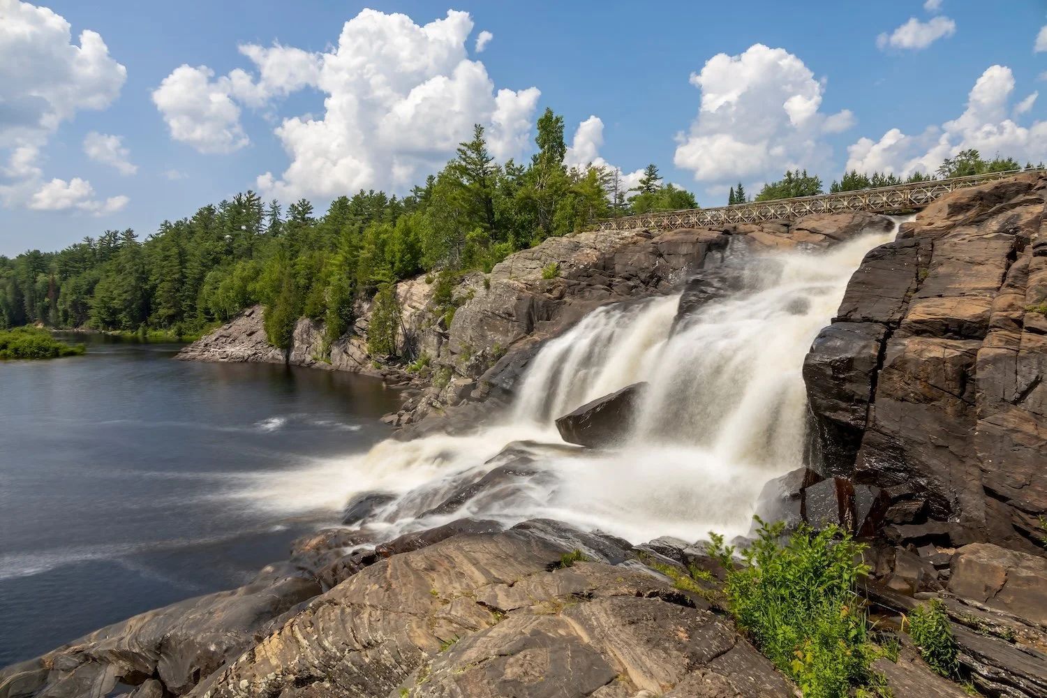 High Falls cascading over rugged Canadian Shield rock into the Muskoka River in Bracebridge, with a footbridge and pine forest in the background