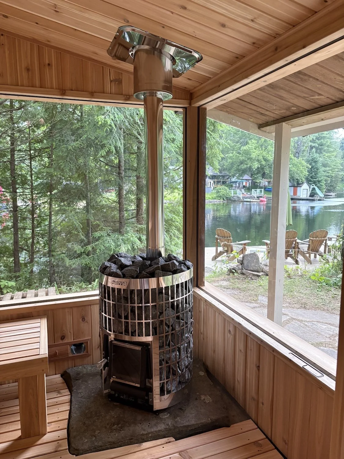 Cedar sauna interior with a wood-burning stove on a stone base and corner windows overlooking the lake and dock