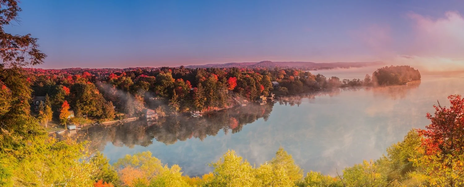 Panoramic autumn view from the Dorset Lookout Tower showing vibrant fall colours, morning mist rising over a calm lake, and cottages nestled among red and orange trees