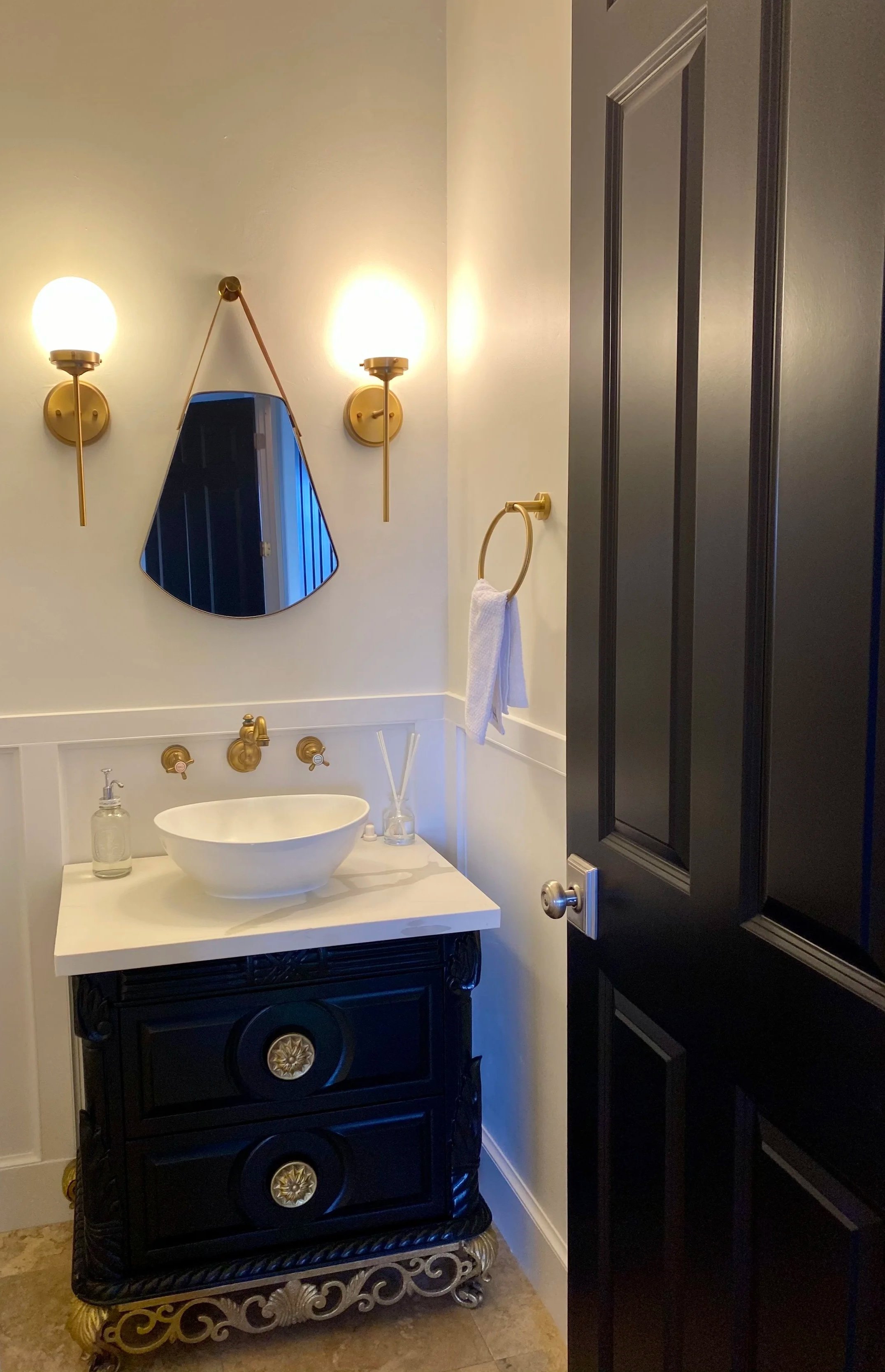 A bathroom featuring a black vintage-style vanity with gold accents, a white vessel sink, two wall-mounted gold light fixtures, a small mirror hanging between the lights, a towel ring with a white towel, and a dark door with a silver doorknob.