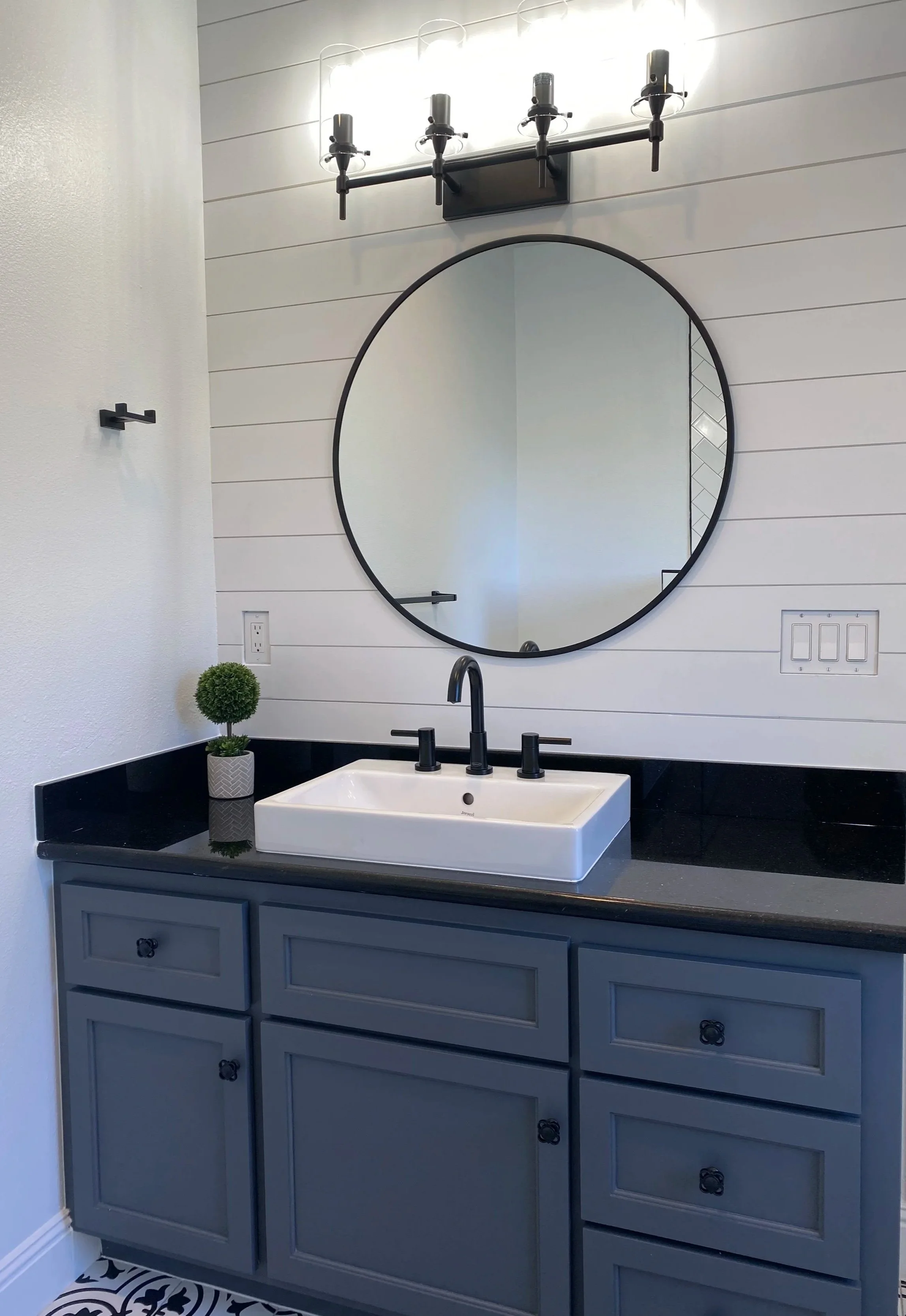 Bathroom vanity with gray cabinets, white rectangular vessel sink, black faucet, round mirror, black light fixture, small potted plant, electrical outlet, and black-and-white patterned floor.