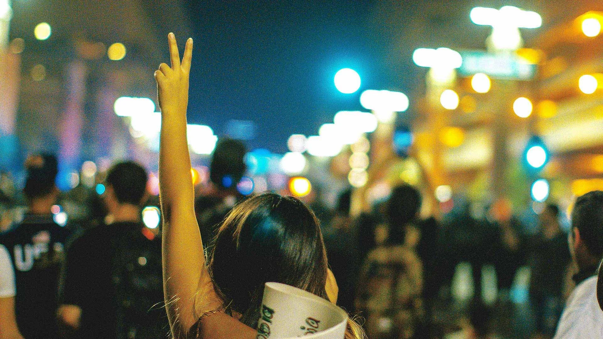 Decorative image of crowd outside at night with woman holding up hand with two fingers "bunny finger" style