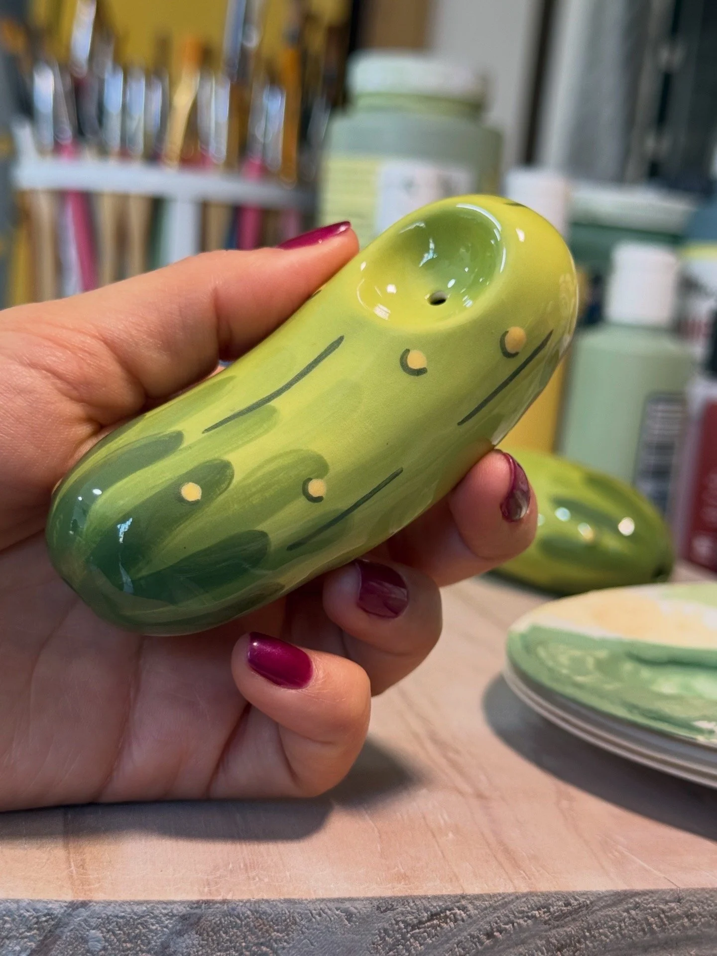 Hand holding a ceramic pickle-shaped ceramic pipe, painted in shades of green and yellow with yellow dots and lines, in a store with shelves of ceramics and other items in the background.