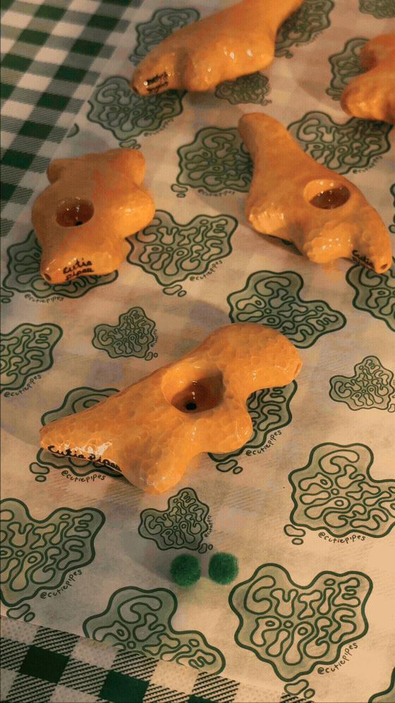 Several dinno nugget ceramic figurines placed on a patterned surface with the @cuteuripes logo and two small green pom-pom balls in front.