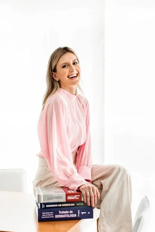 Dra. Paola Mattos - mulher sorridente com cabelo loiro, usando blusa rosa e calça bege, sentada sobre livros empilhados, ao lado de uma mesa branca, em ambiente bem iluminado.