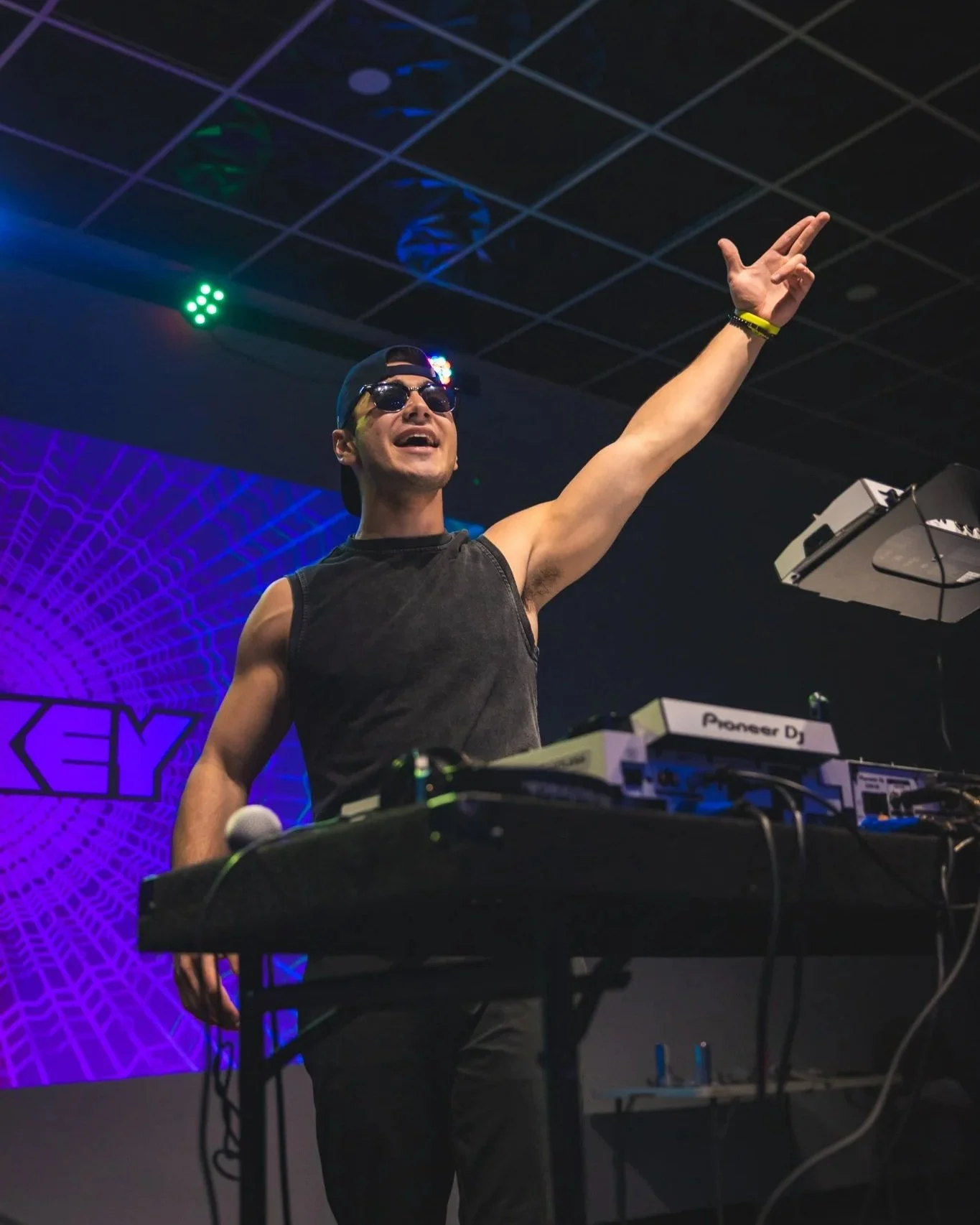 DJ Turkey wearing sunglasses, a backward cap, and a sleeveless shirt, is performing at a music event. He is standing behind DJ equipment and raising his right hand in the air. The background features colorful lights and a textured graphic design.