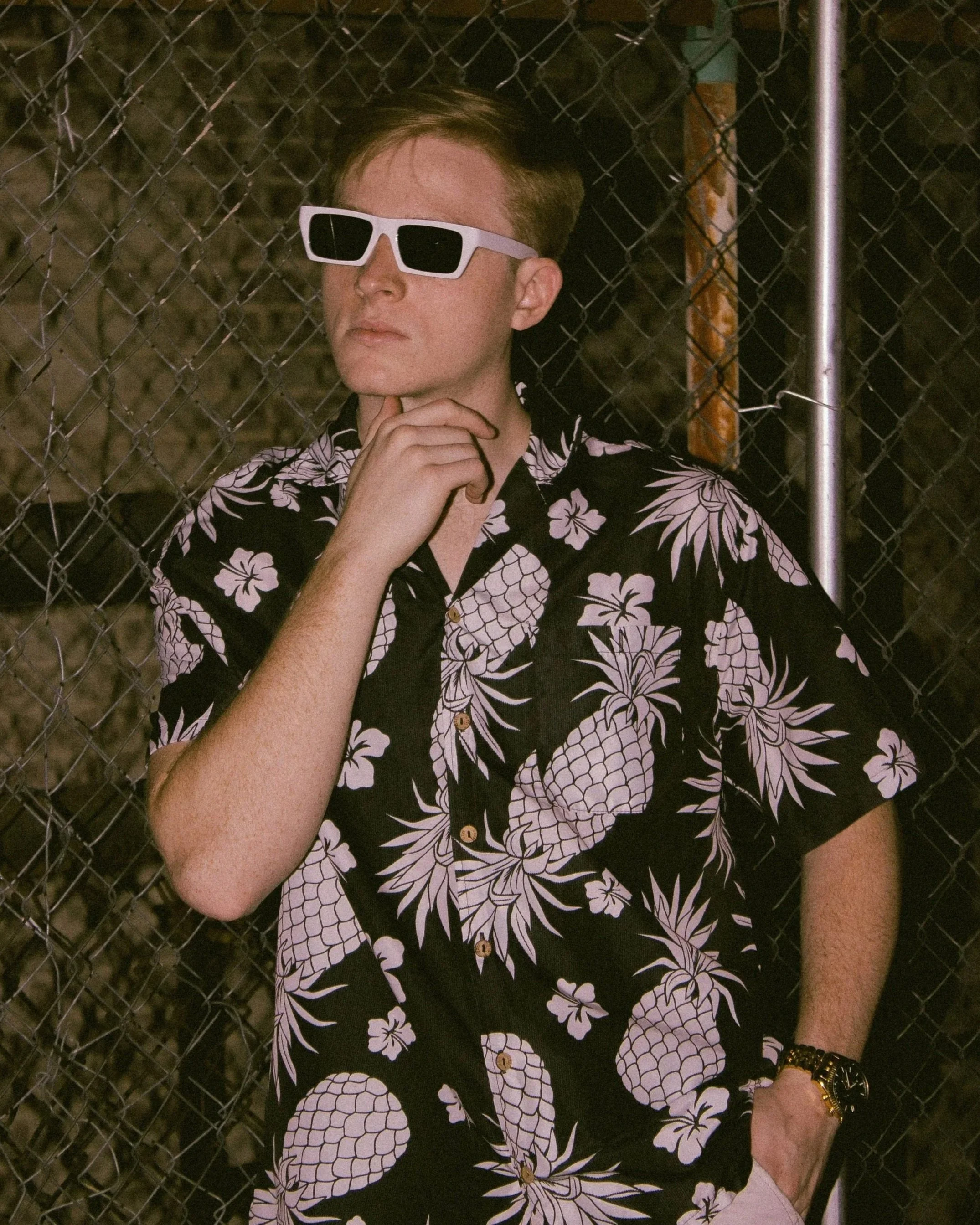 DJ Wyder, a blond man with blond hair and light skin wearing large white sunglasses and a black Hawaiian shirt with white floral patterns, standing against a chain-link fence at night, posing for a headshot.