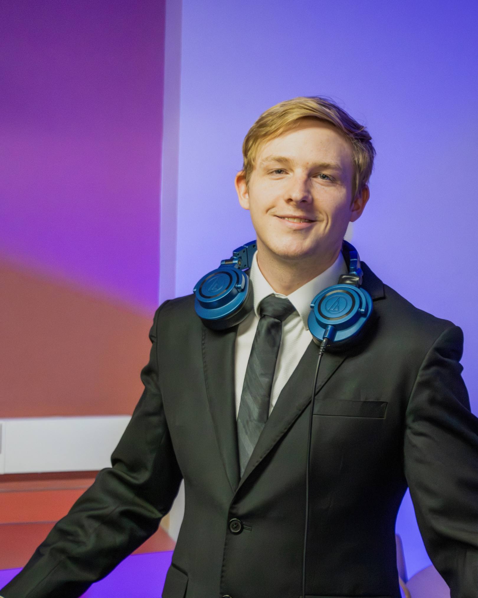 DJ Wyder wearing a black suit, white shirt, and black tie, with large blue headphones around his neck, smiling in front of a colorful gradient background with shades of purple, blue, and red.