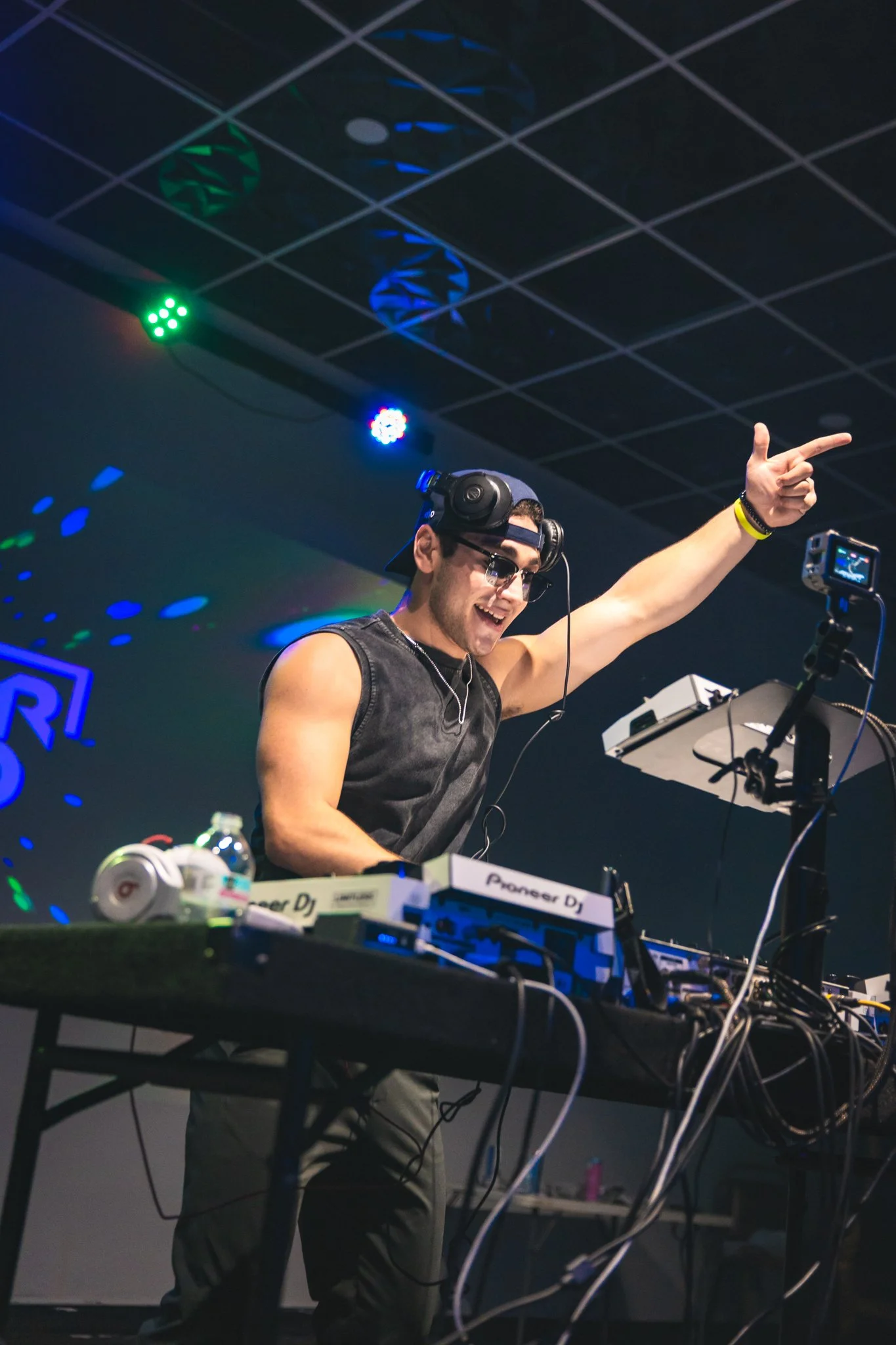 DJ Turkey is wearing a black sleeveless shirt, glasses, and headphones, and smiling while pointing with his right hand. He is next to a DJ setup with turntables and other equipment, in a dark room with colorful lights on the ceiling.