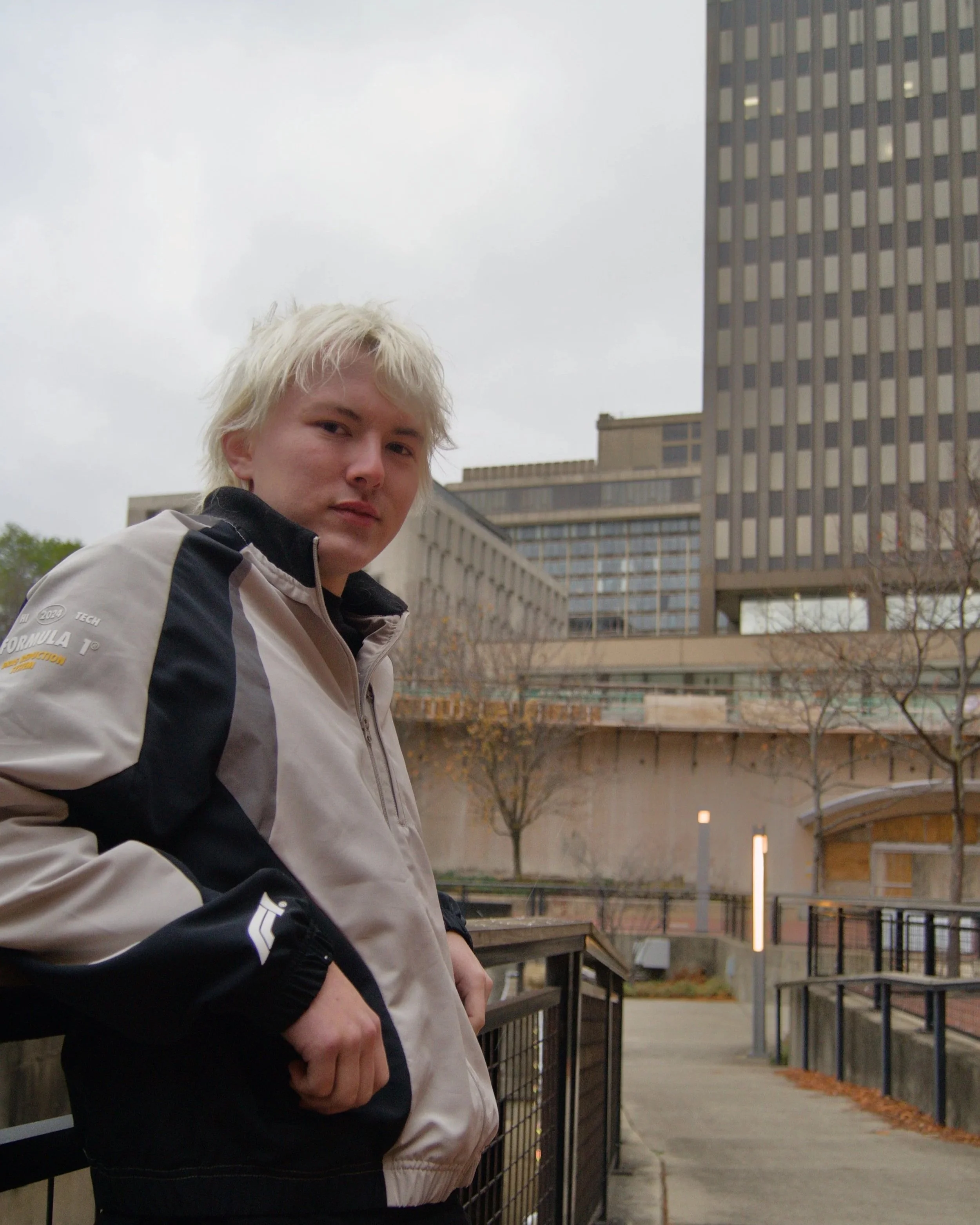 DJ Draco (Matt Miller), with platinum blonde hair wearing a beige and black jacket standing outdoors near a metal railing in an urban area with high-rise buildings and trees in the background.
