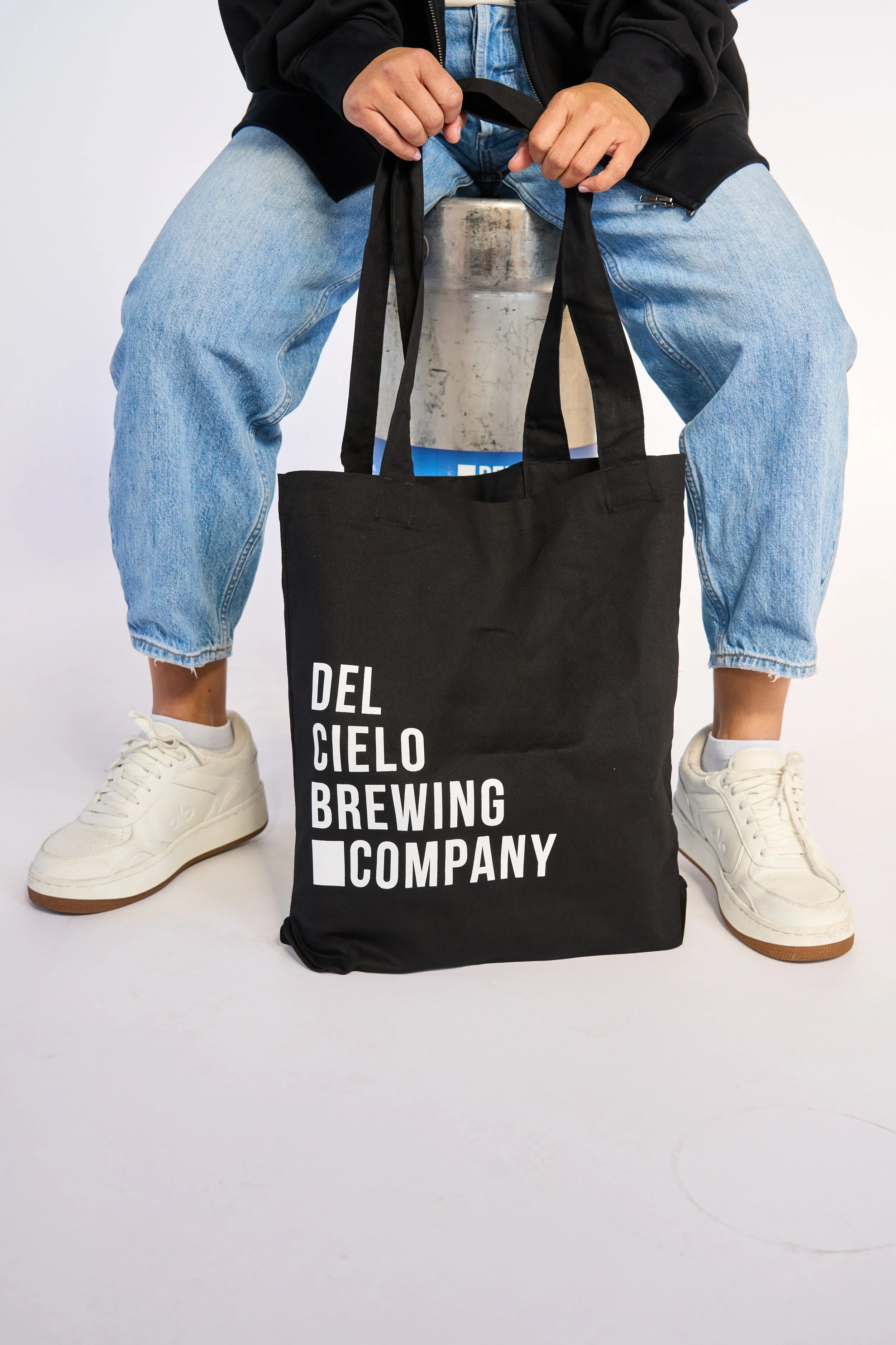 A person sitting on a metal keg, wearing a black jacket, light blue jeans, and white sneakers, holding a black tote bag with white text, in front of a plain white background.