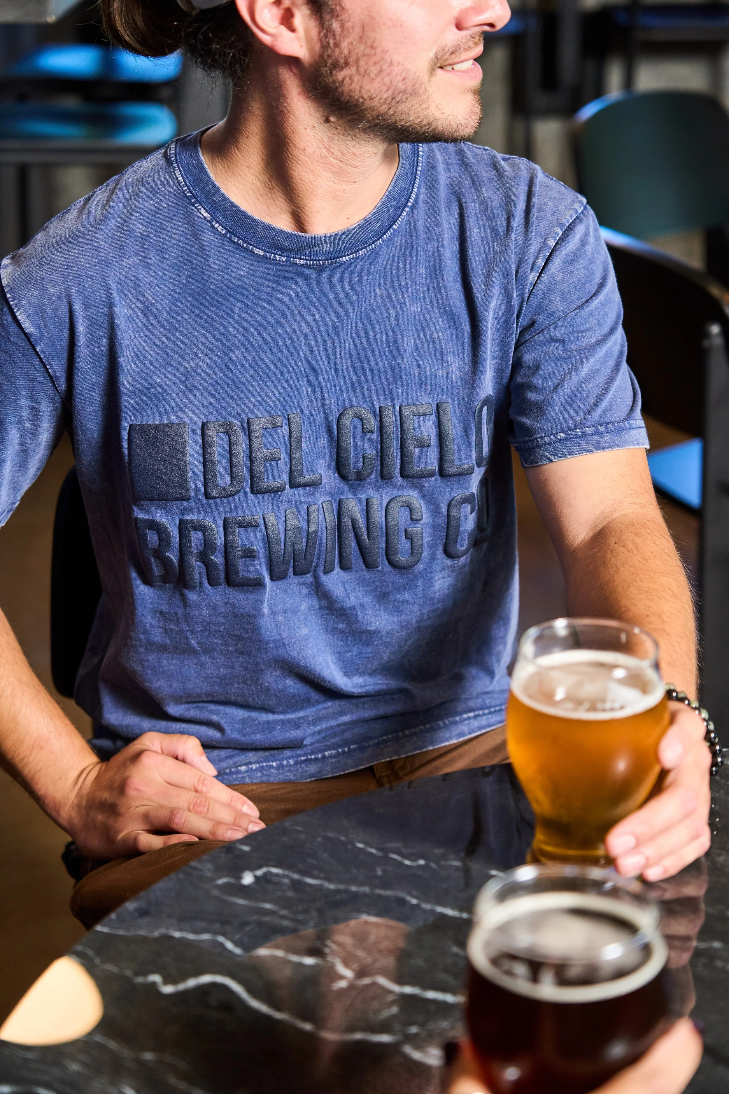 A man wearing a blue t-shirt that reads "Del Cielo Brewing Co" is holding a glass of beer. The background shows chairs and a bar or pub setting.