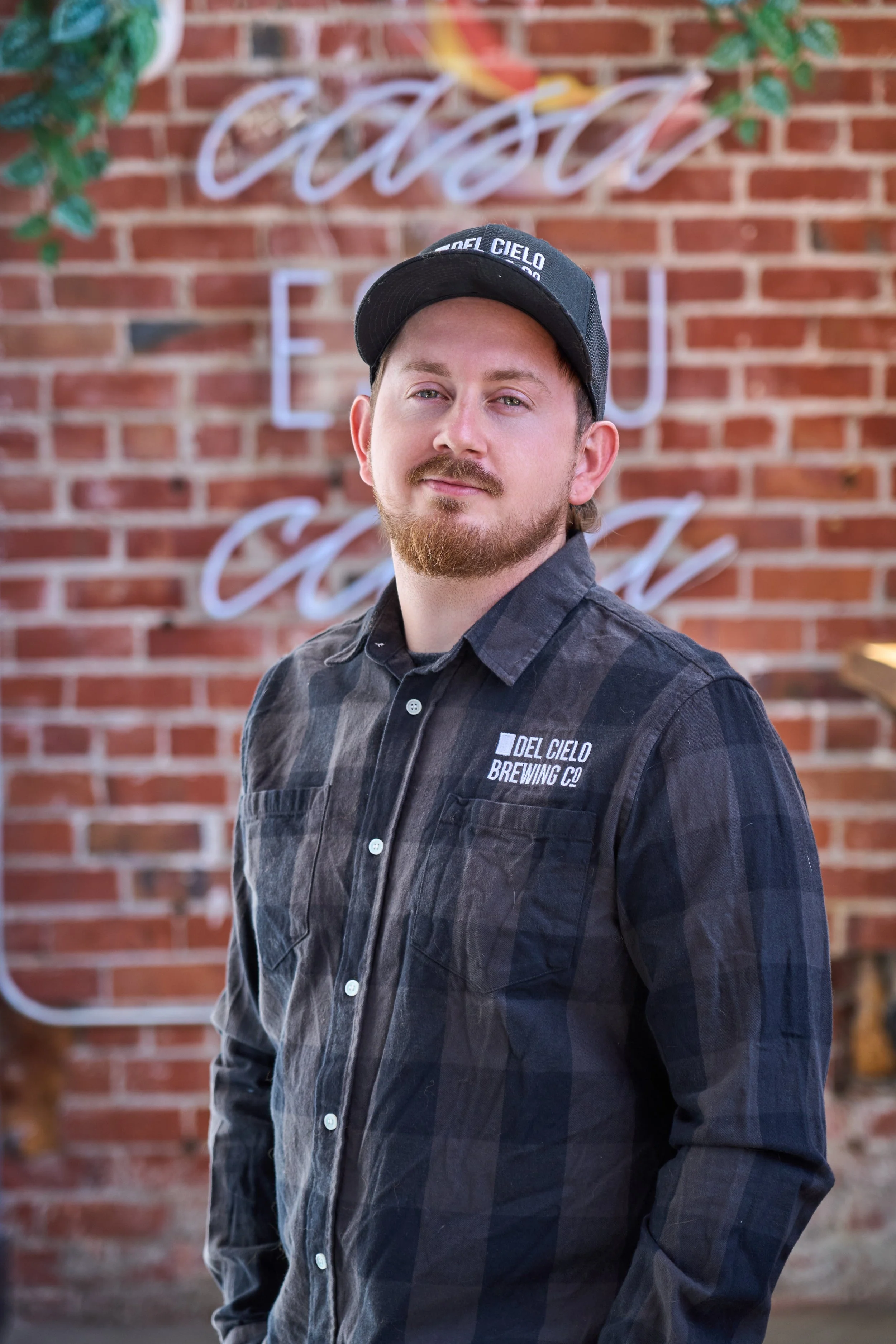 A man with light skin, a beard, and mustache wearing a black cap with logo and a dark plaid shirt, standing in front of a brick wall with a neon sign that reads 'Casa' in cursive, partially visible.