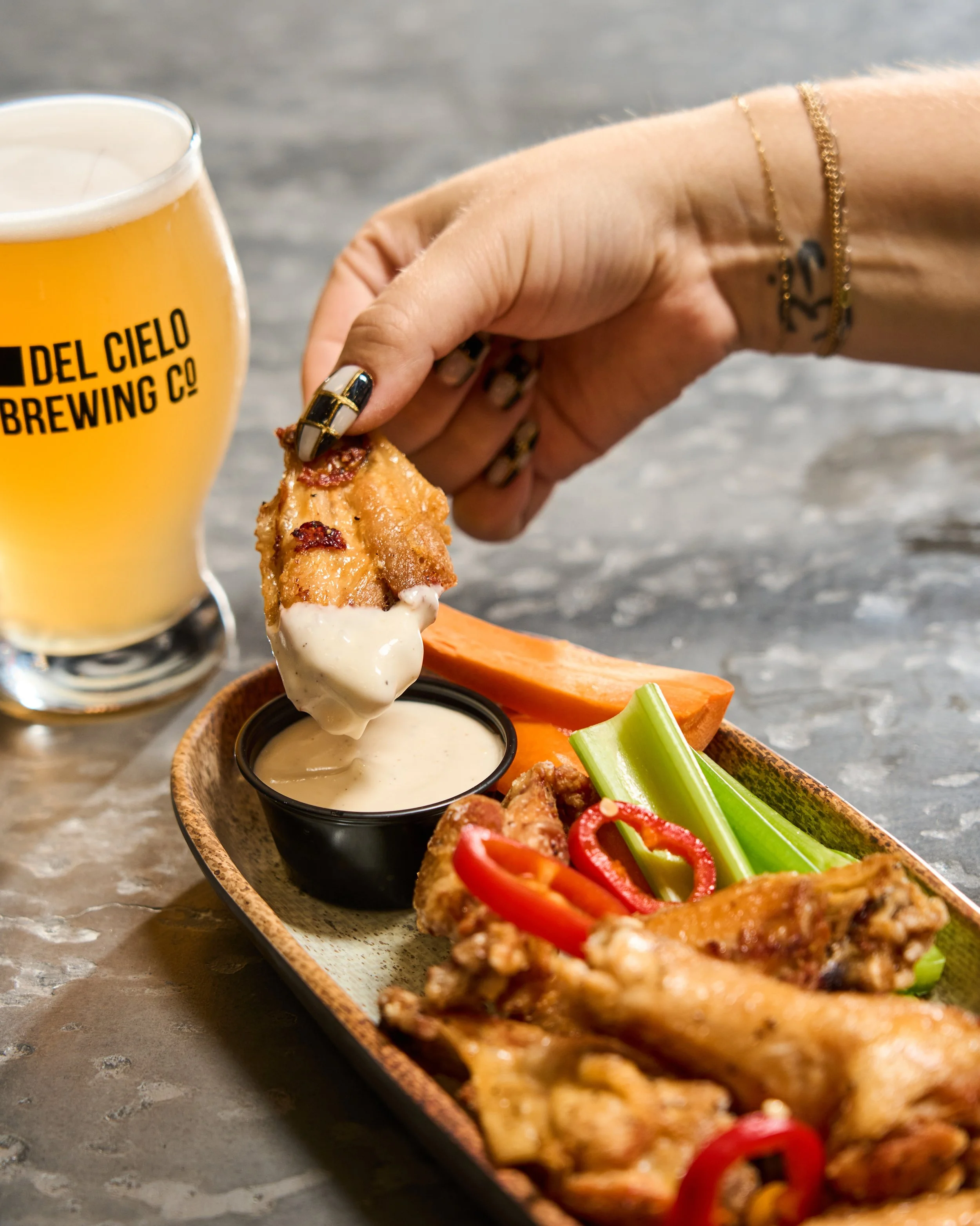 A hand with decorated nails holding a piece of cooked chicken that has white sauce. A beer in a glass labeled 'Del Cielo Brewing Co' is in the background, and a wooden tray holds more chicken, celery sticks, red pepper slices, and dipping sauce.