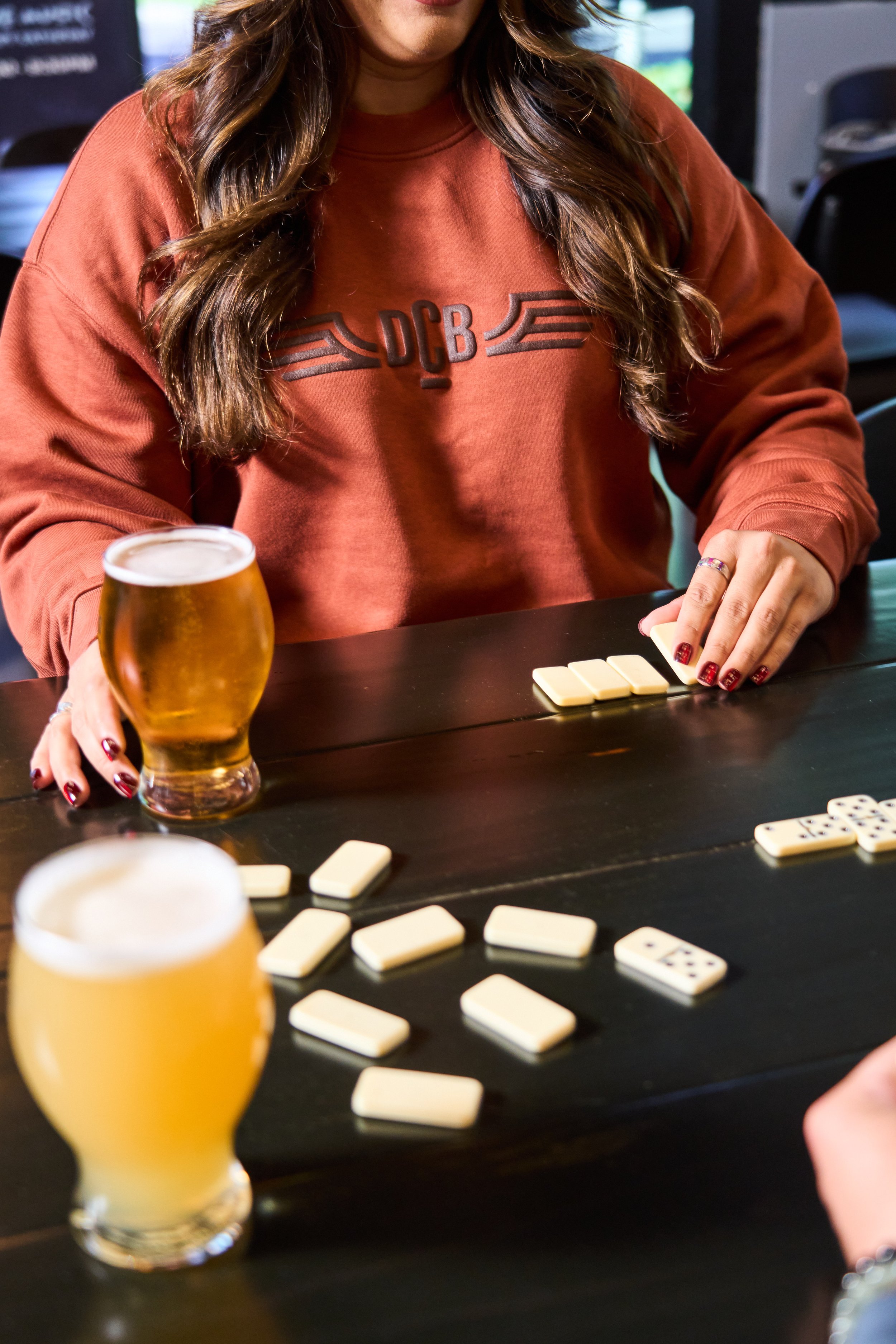 A woman with long, wavy brown hair wearing a rust sweatshirt playing dominoes at a dark-colored table. There is a glass of beer in the foreground and several domino tiles on the table.