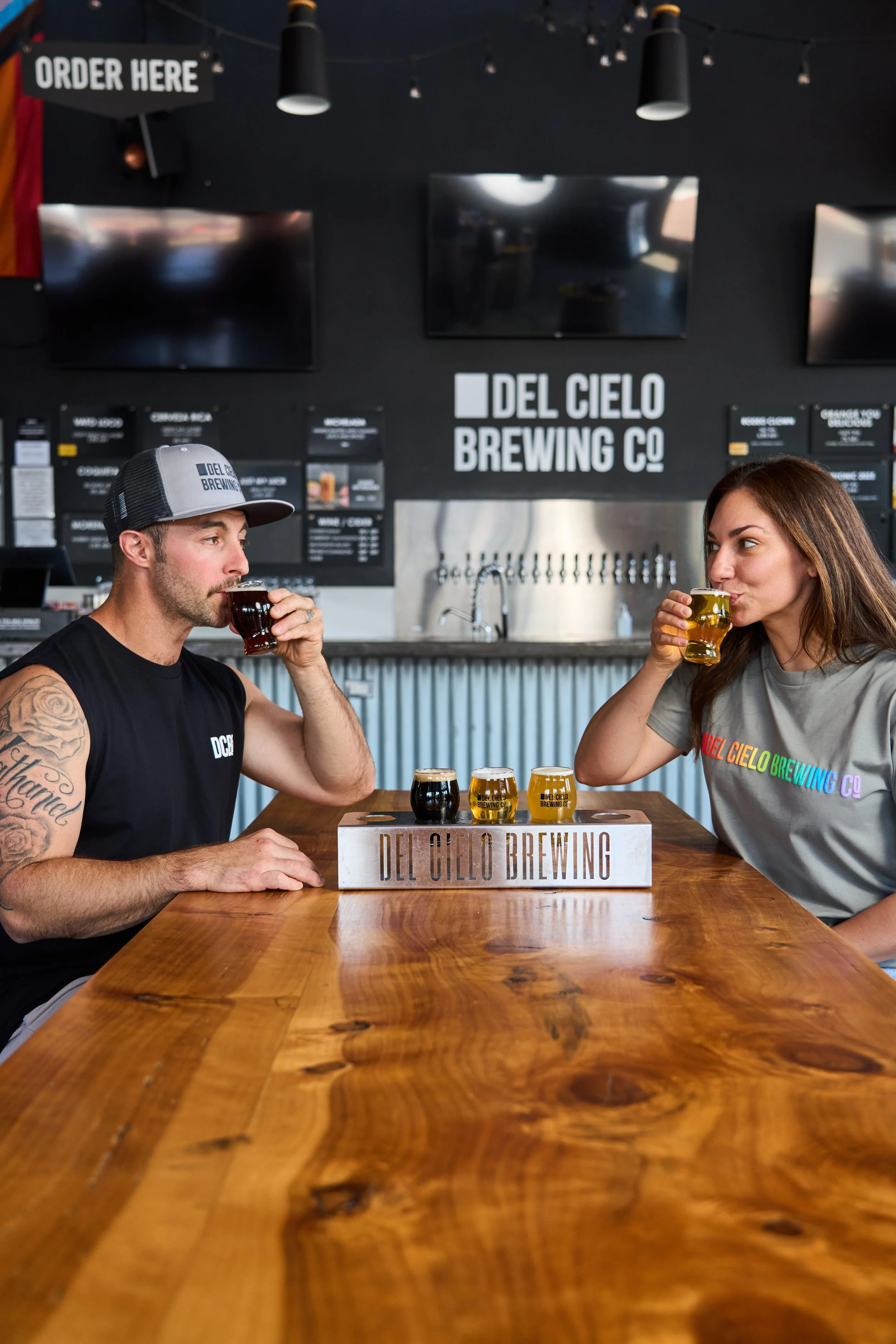 A man and a woman sitting at a wooden table, each drinking a small glass of beer inside Del Cielo Brewing Co. The man is wearing a gray baseball cap and sleeveless shirt, and the woman has long brown hair and is wearing a gray T-shirt. The background