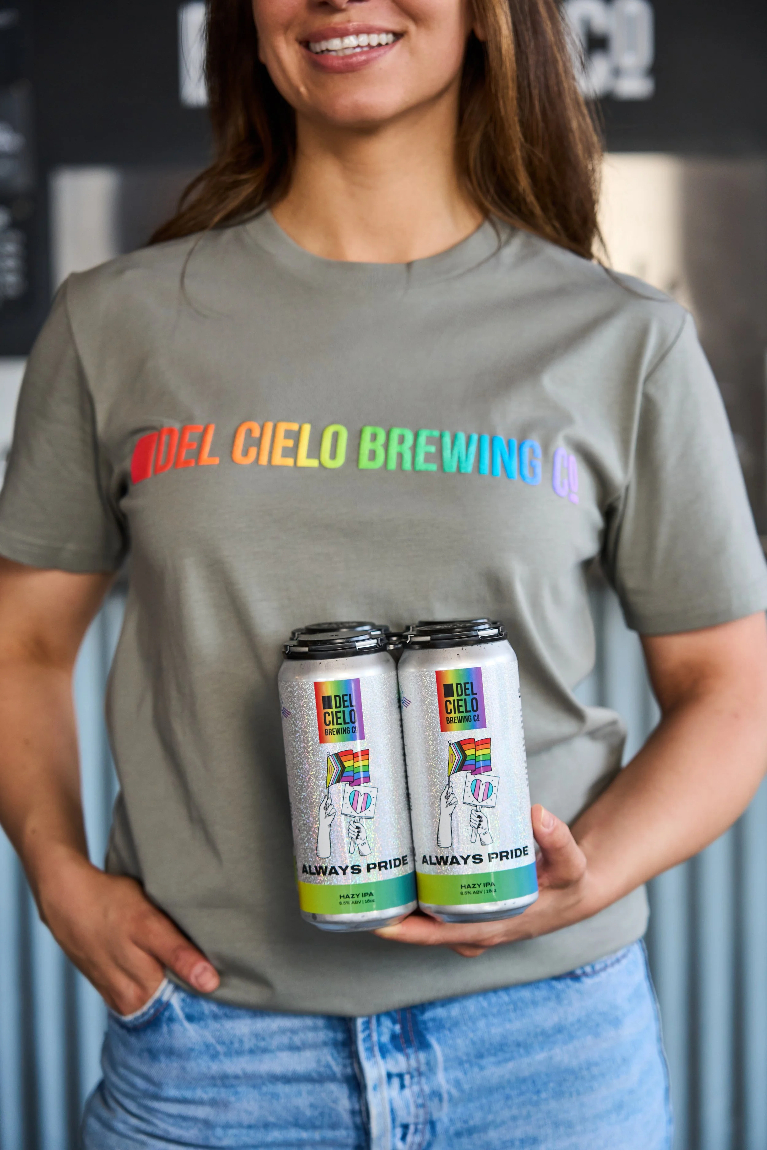 A person wearing a gray 'Del Cielo Brewing Co' T-shirt, holding two cans of beer with rainbow and pride-themed labels, standing indoors.
