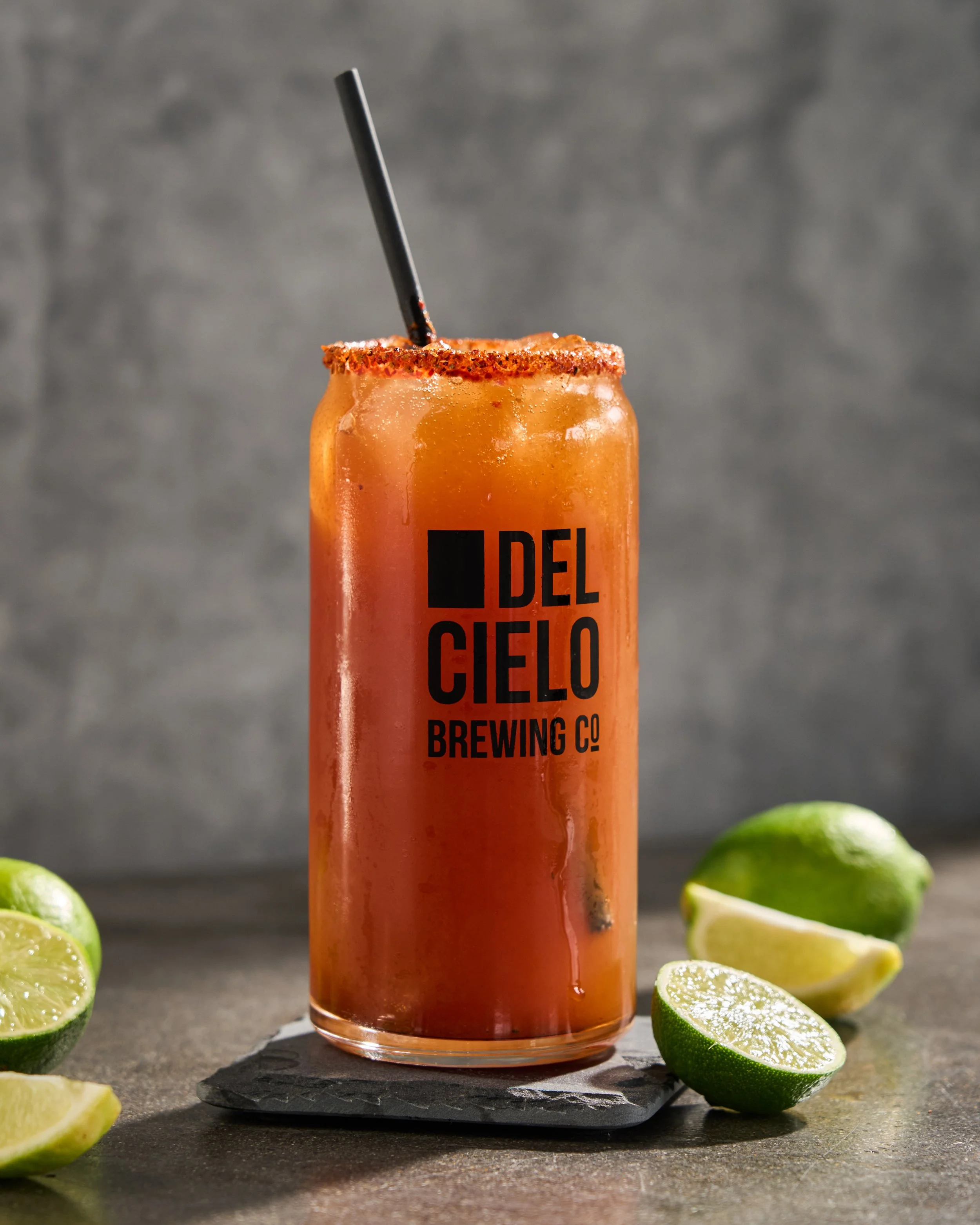 A can of Del Cielo Brewing Co. beverage with a reddish color, garnished with a chili powder rim and a black straw, placed on a black slate coaster on a grey surface, with lime wedges and lime slices around it.