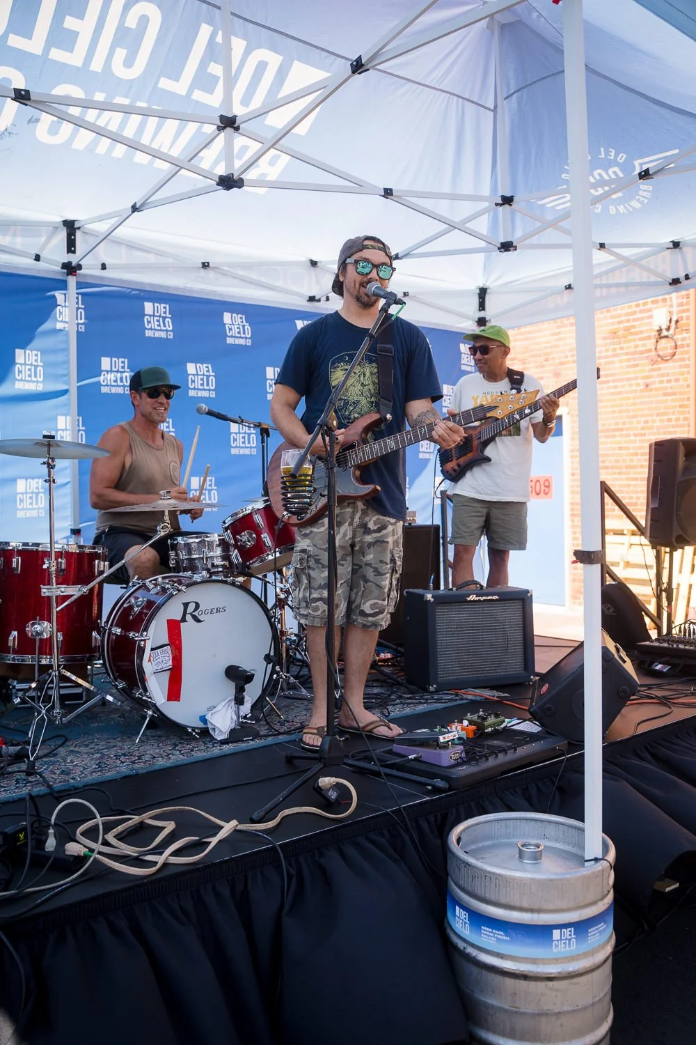 A band of three musicians performing on an outdoor stage under a white canopy, with a blue backdrop that reads 'Del Cielo Brewing Co'. The drummer is wearing sunglasses and a tank top, the bassist is in sunglasses and a white shirt, and the guitarist is in sunglasses, a cap, and camouflage shorts.