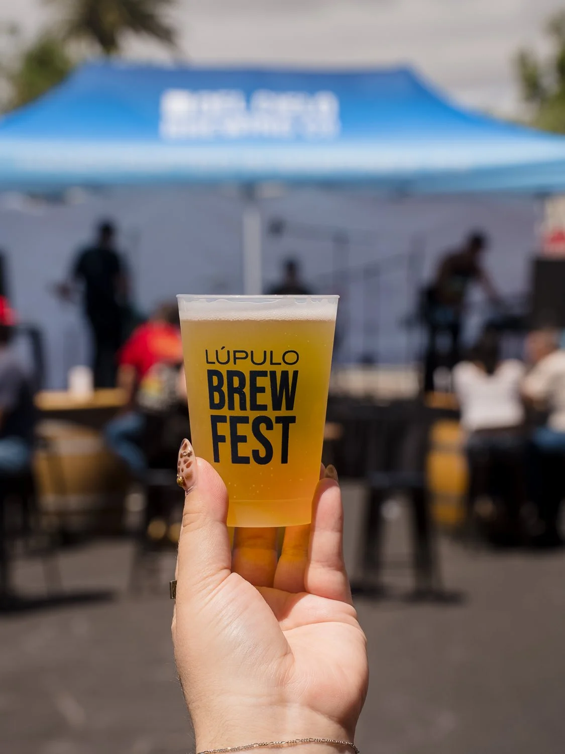 A hand holding a clear plastic cup with a pale yellow beer labeled "Lúpulo Brew Fest" at an outdoor event with a blue canopy and blurred people in the background.