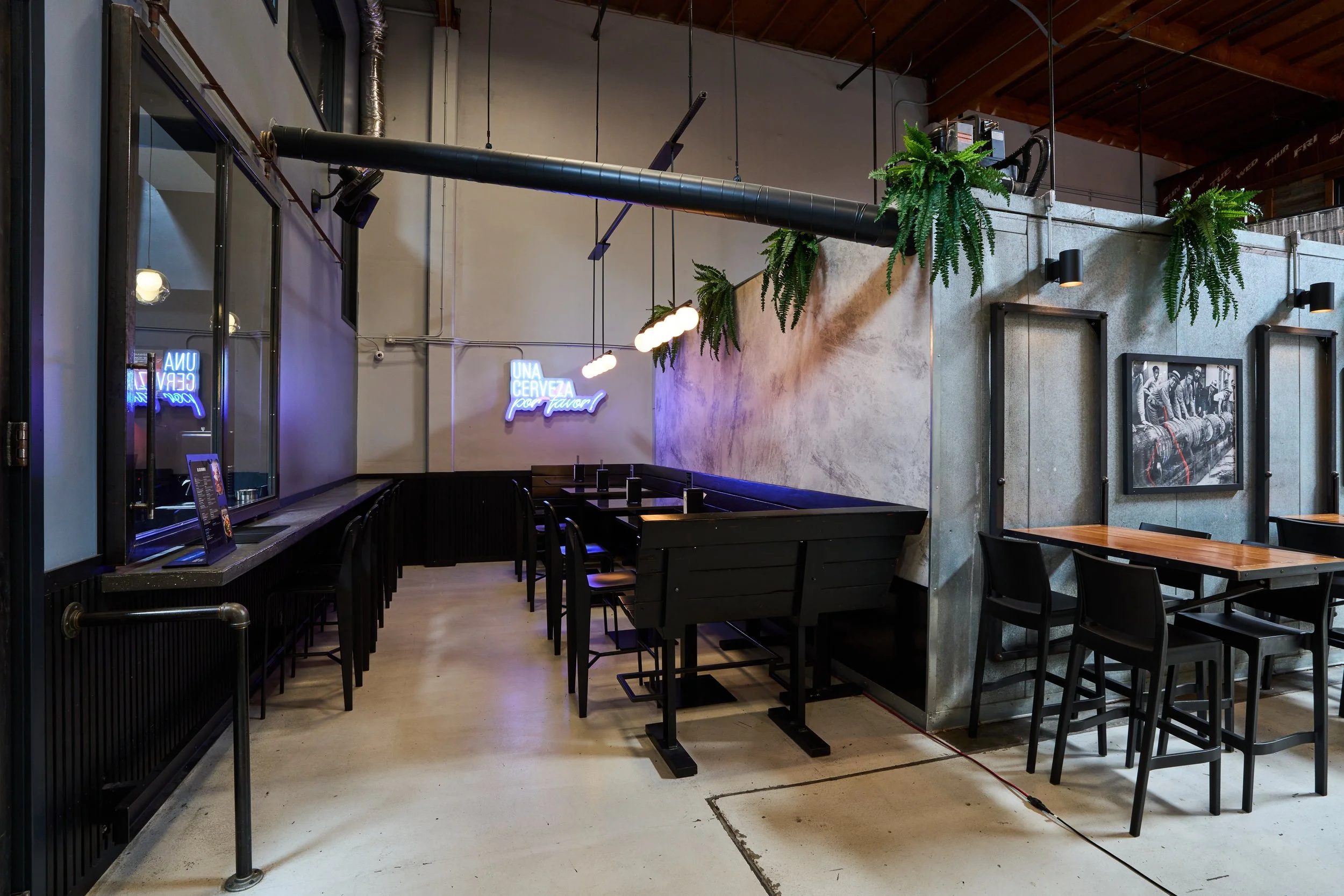 Interior of a modern restaurant with black tables and chairs, hanging plants, framed black and white photo, neon signs, and industrial style decor.