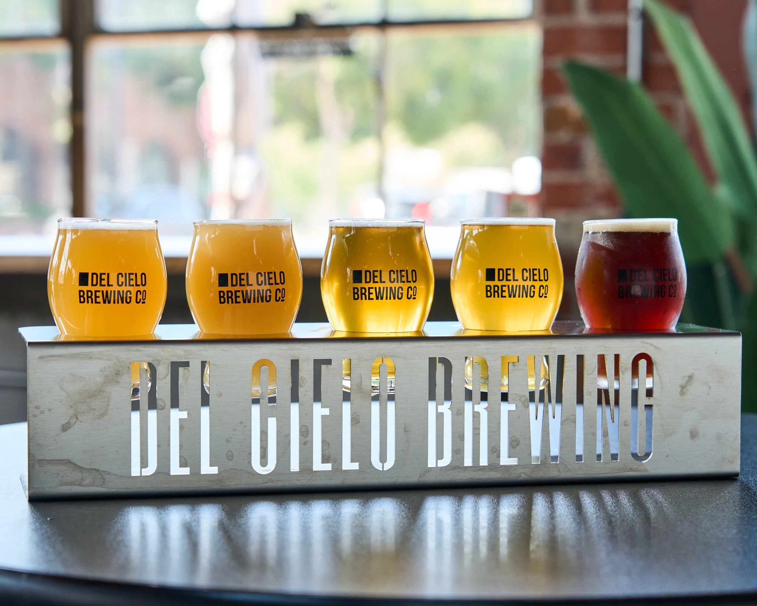 A flight of five colorful beers from Del Cielo Brewing Co on a wooden tray with the brewery's name cut out, set on a table in front of a window with blurred outdoor scenery.