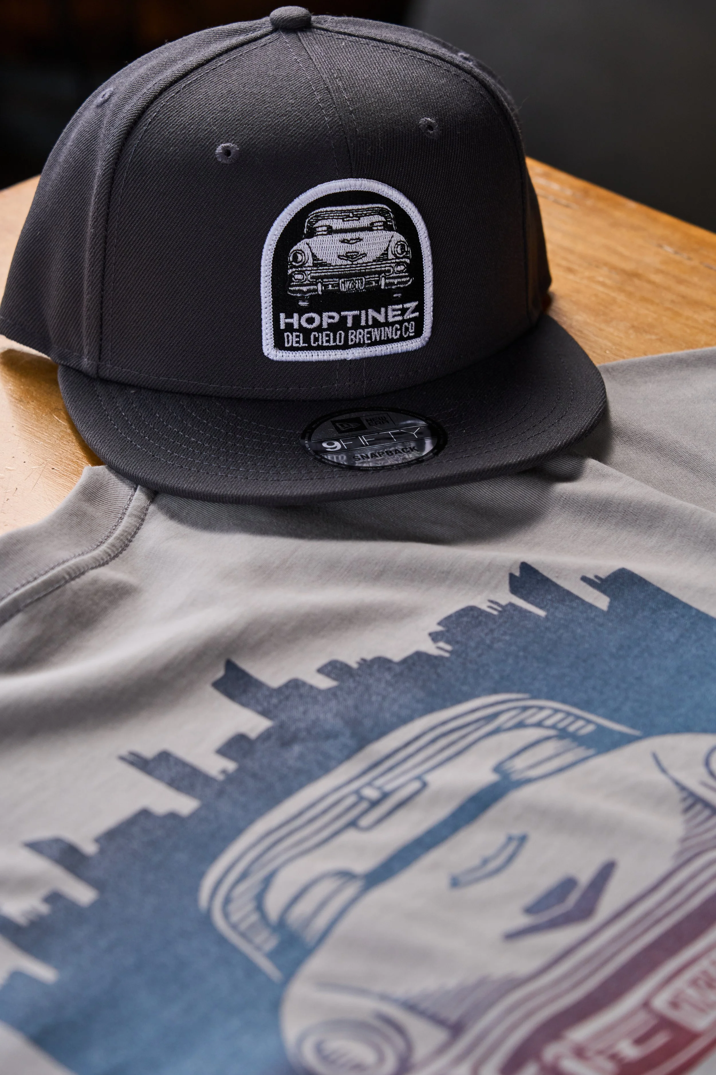 Black baseball cap with embroidered patch featuring a vintage car and text 'HOPTINEZ DEL CIELO BREWING CO', resting on a gray t-shirt on a wooden surface.