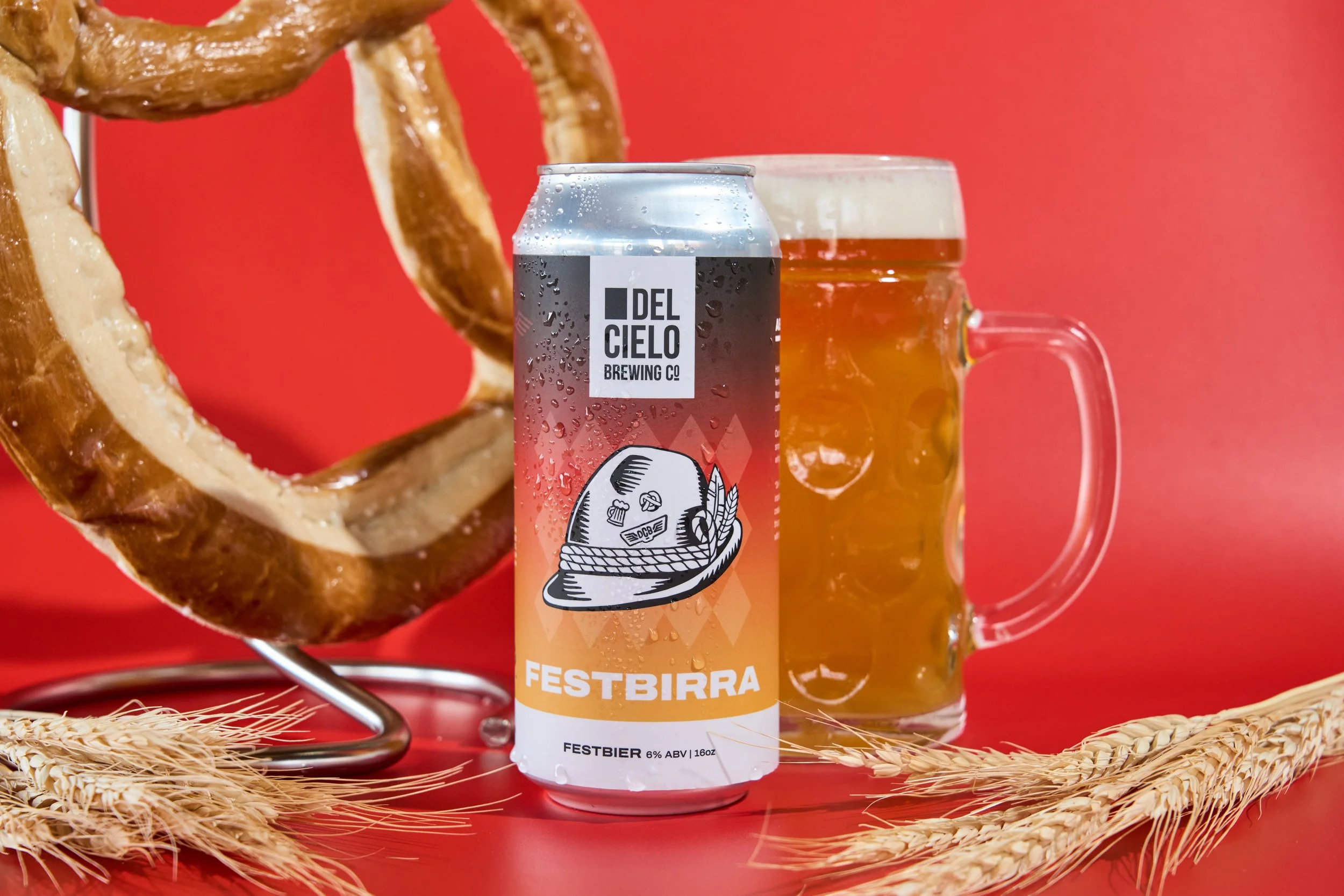 A can of Festbier from Del Cielo Brewing Co, a glass mug filled with beer, a pretzel, and wheat stalks on a red background.
