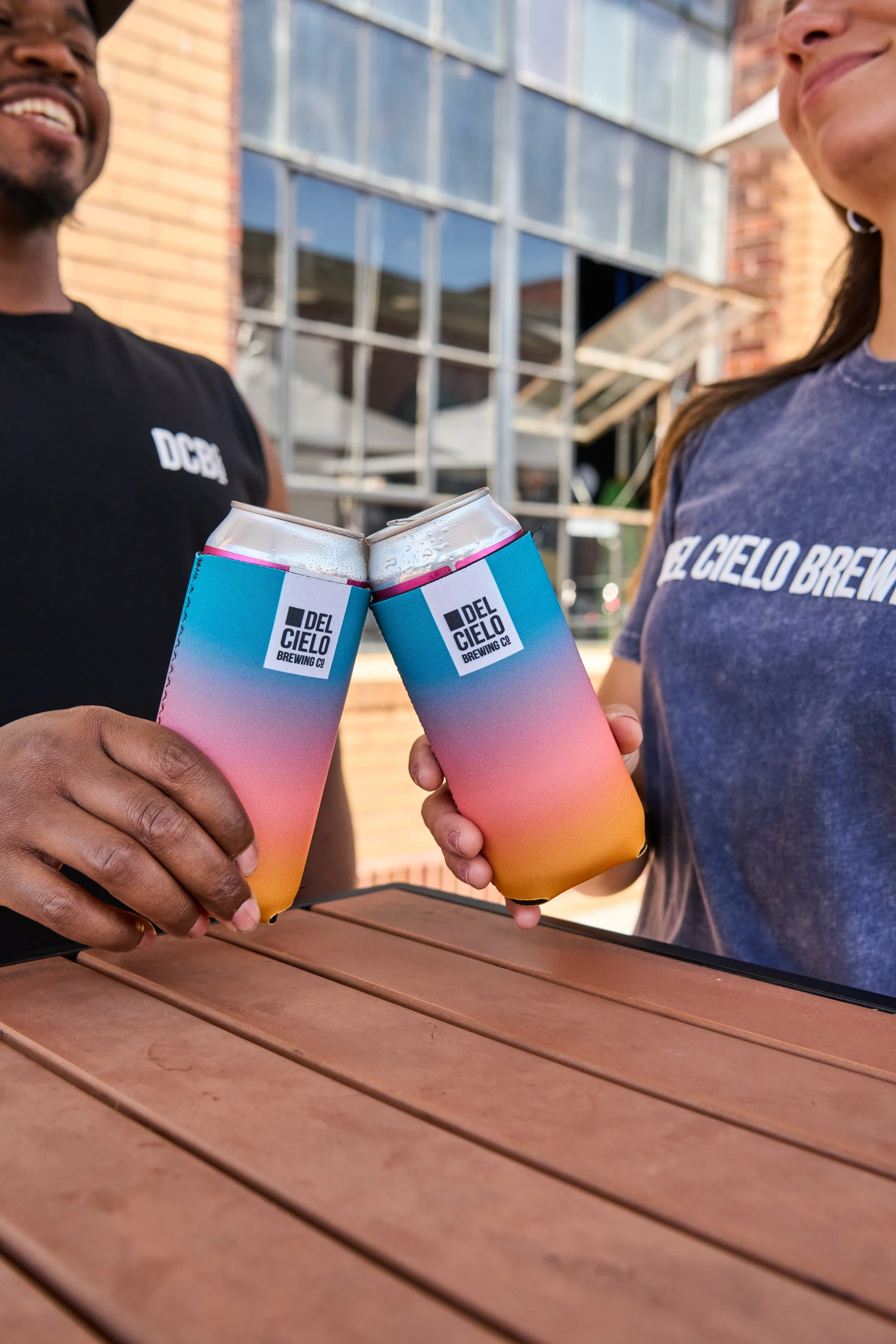 Two people are clinking cans of Del Cielo Brewing Co. beer outdoors, with a brick building and large windows in the background. One person is wearing a black shirt and the other a blue shirt.