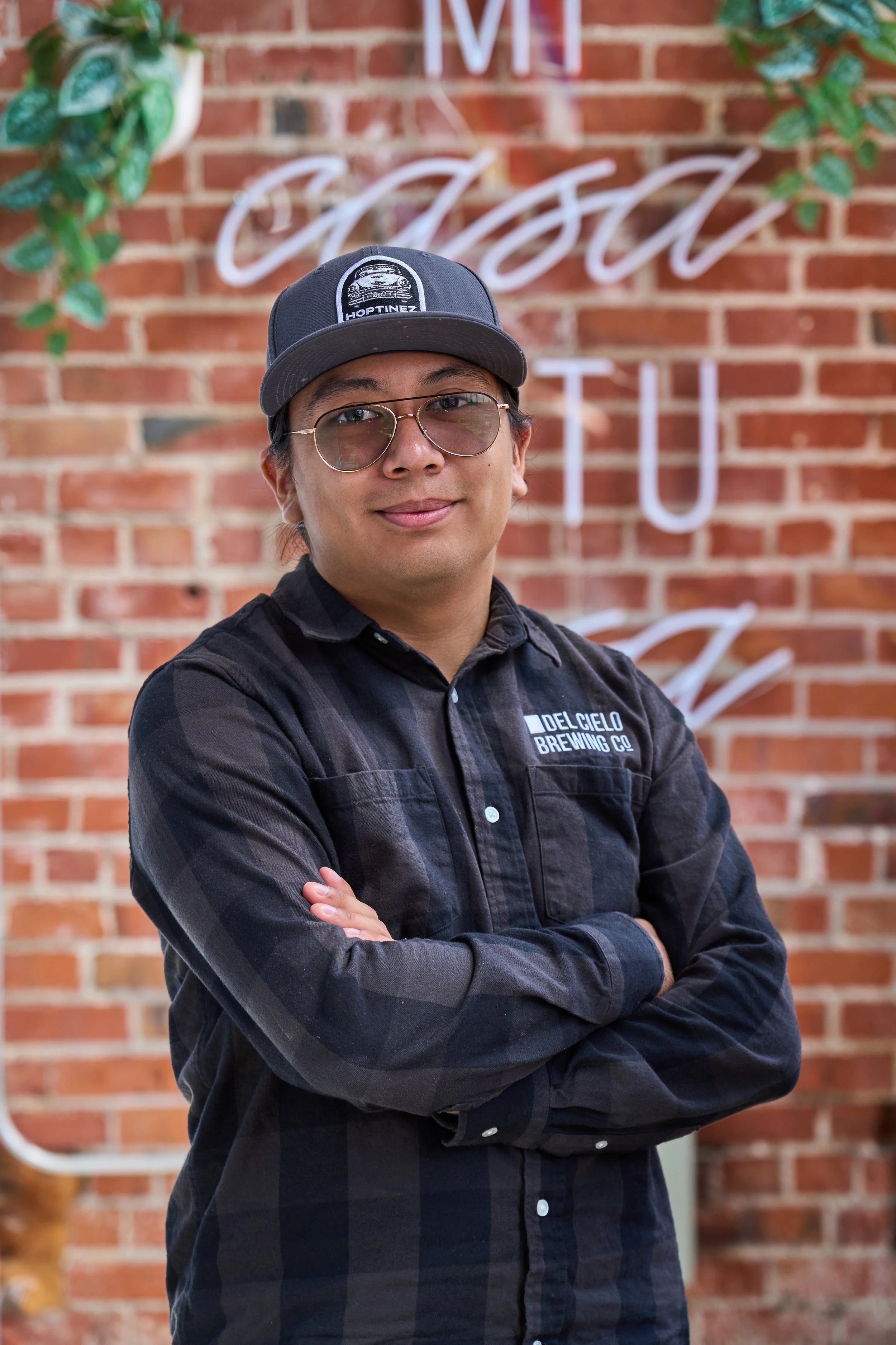 Man wearing glasses and a black shirt with crossed arms standing in front of a brick wall with neon sign reading 'Casa' and greenery on the sides.