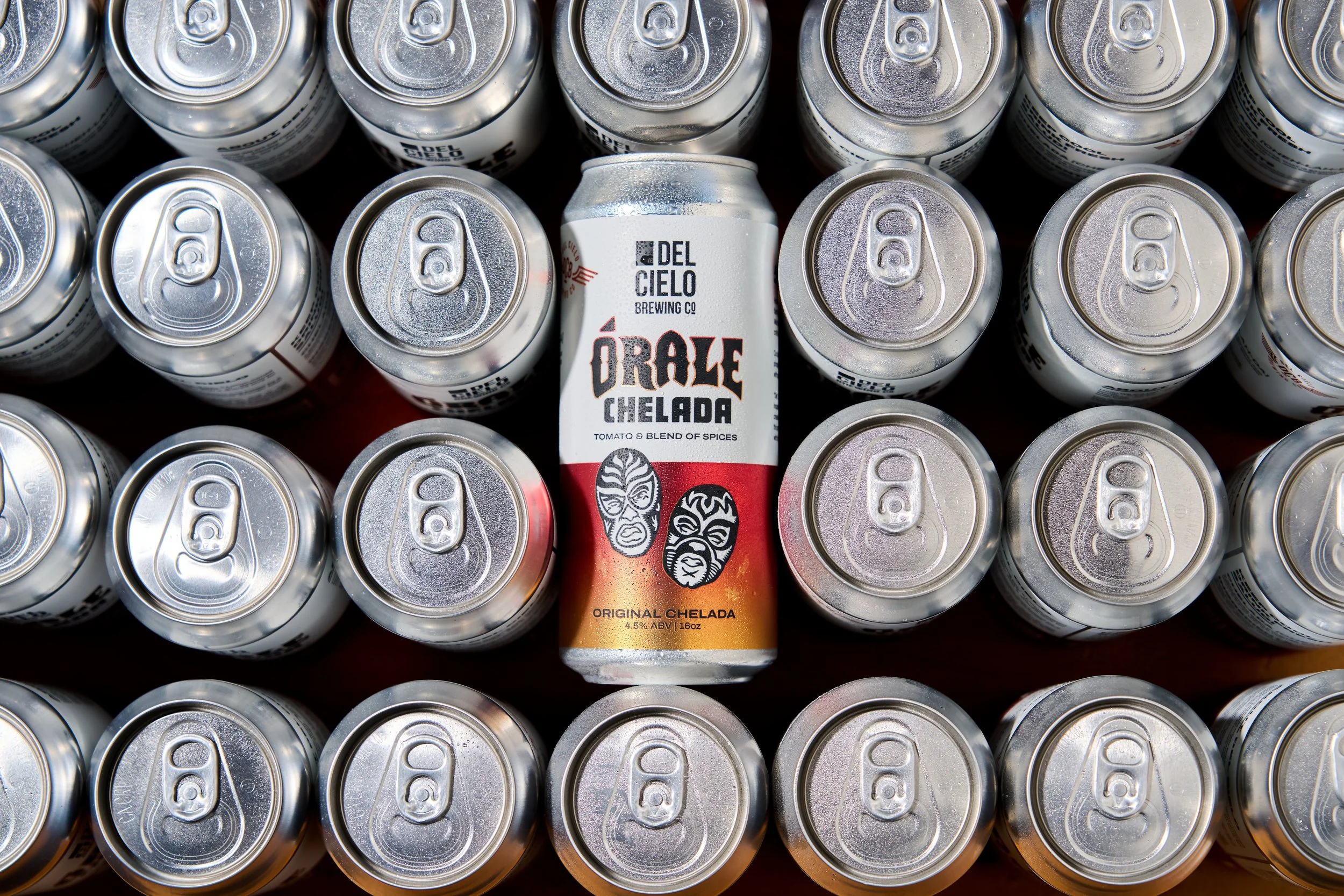 A collection of silver cans of chelada with a single can in the center labeled 'Del Cielo Brewing Co.' with orange and white colors, surrounded by other similar unbranded silver cans.
