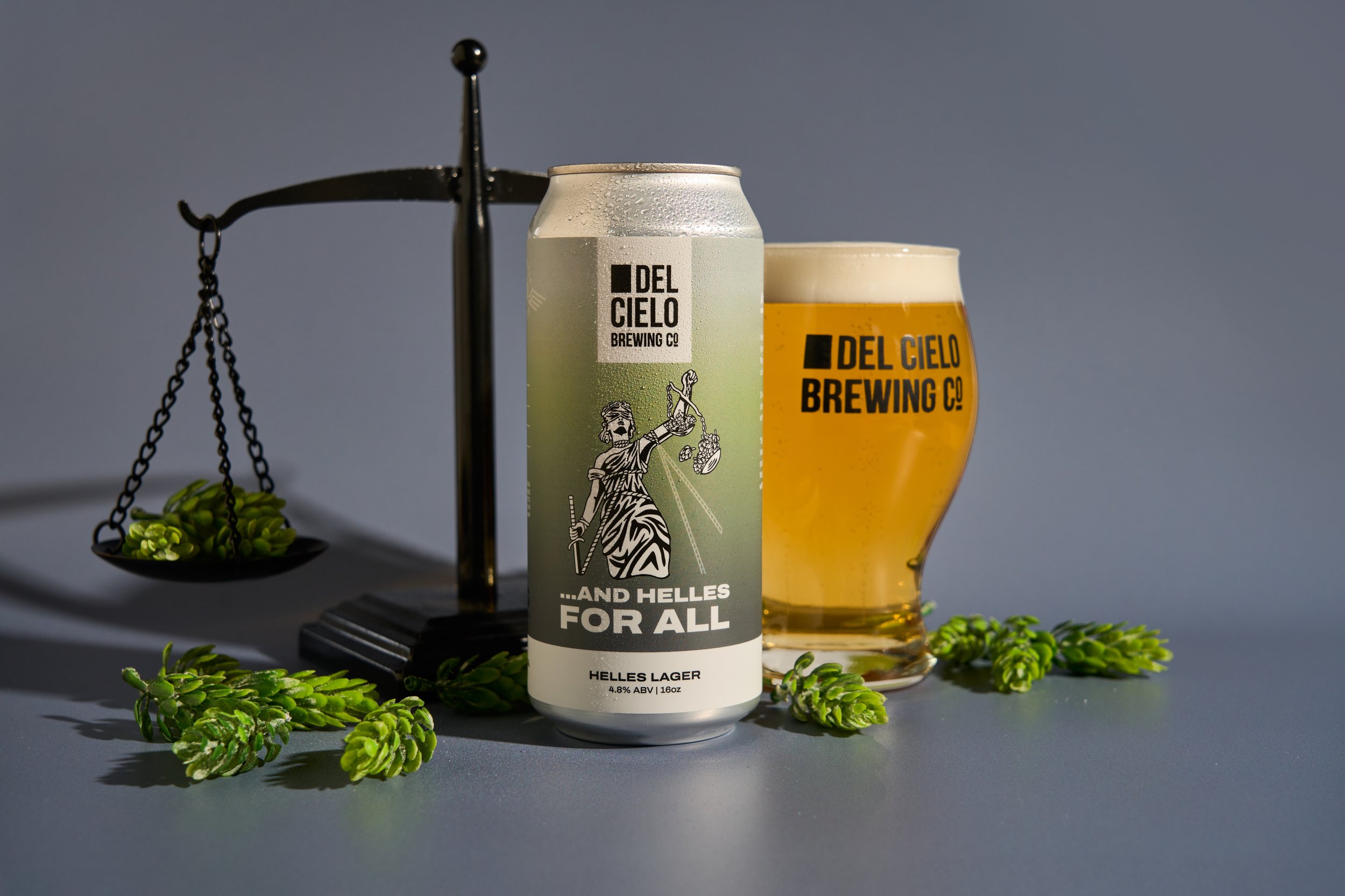 A can of Del Cielo brewing co. Helles Lager beer with a glass of beer beside it, placed on a surface with green decorative plants around. A balance scale is partially visible in the background.