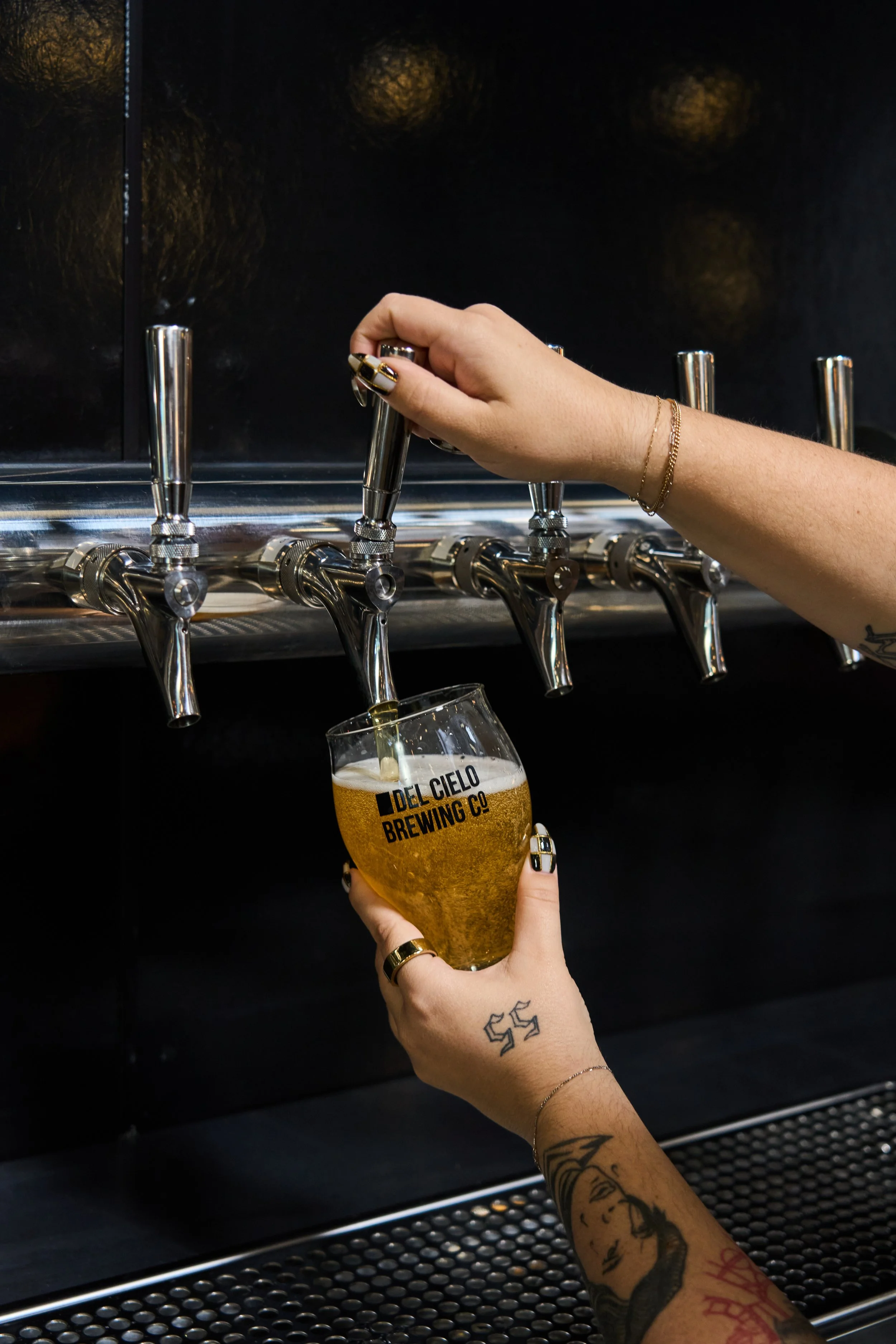 A person with tattoos on their arm pouring beer from a tap at a brewery, holding a glass with the logo of Idel Cielo Brewing Co.