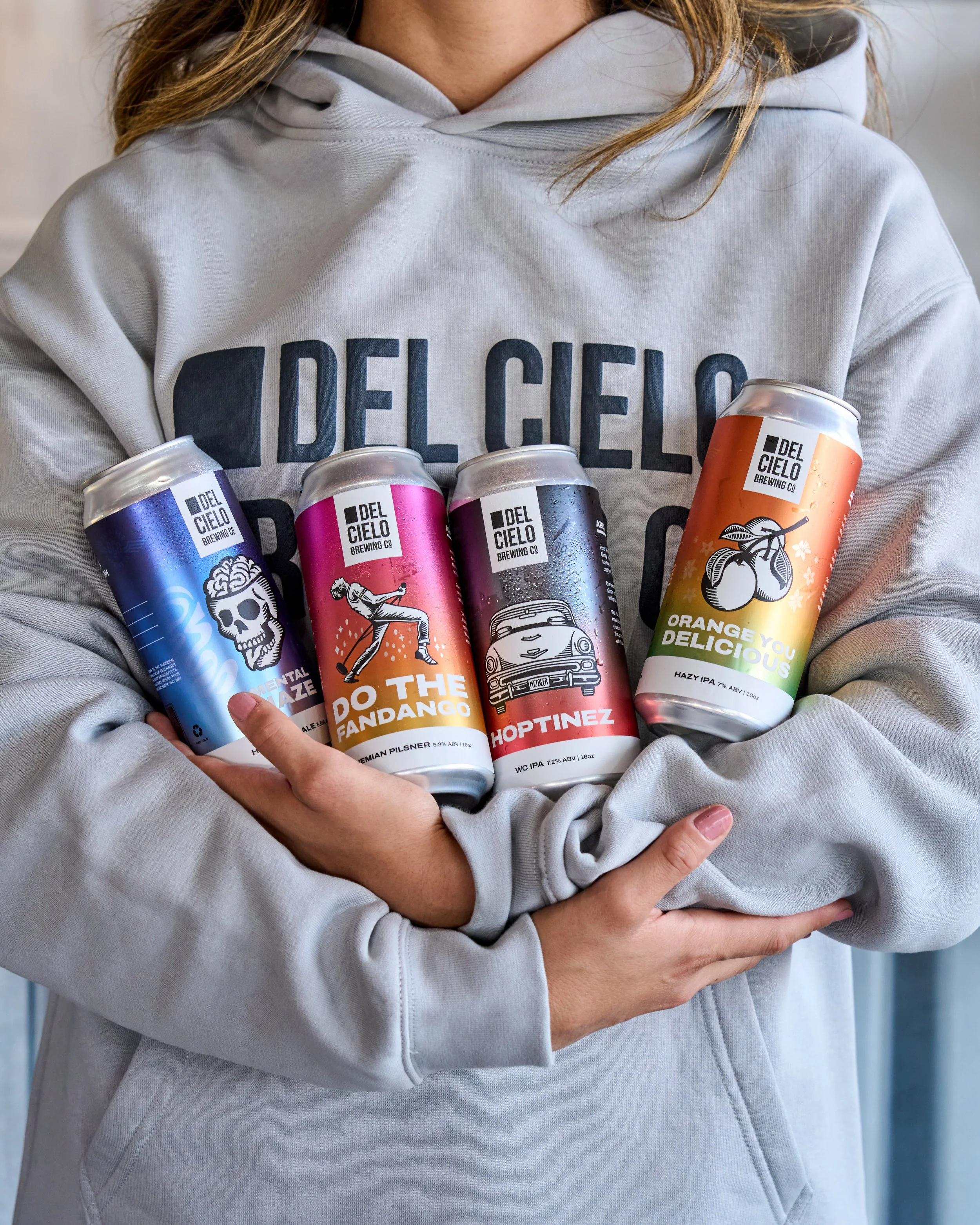 A person wearing a light gray hoodie and holding four cans of beer from Del Cielo Brewing Co. against a neutral background.