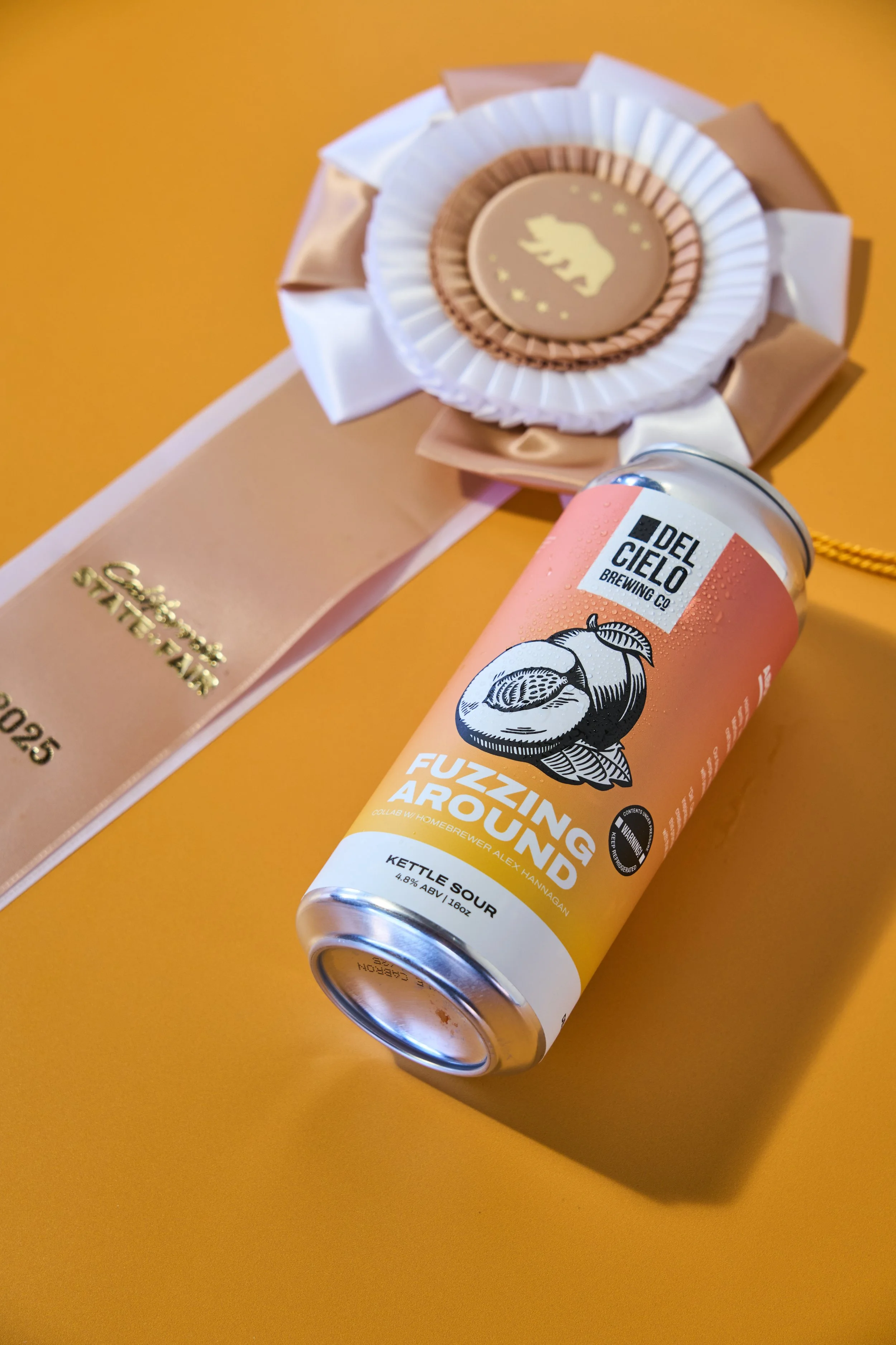 A can of Del Cielo Brewing Co.'s Fuzzing Around kettle sour beer with an illustrations of a peach, placed on a yellow surface next to a pink ribbon award and a beige award ribbon.
