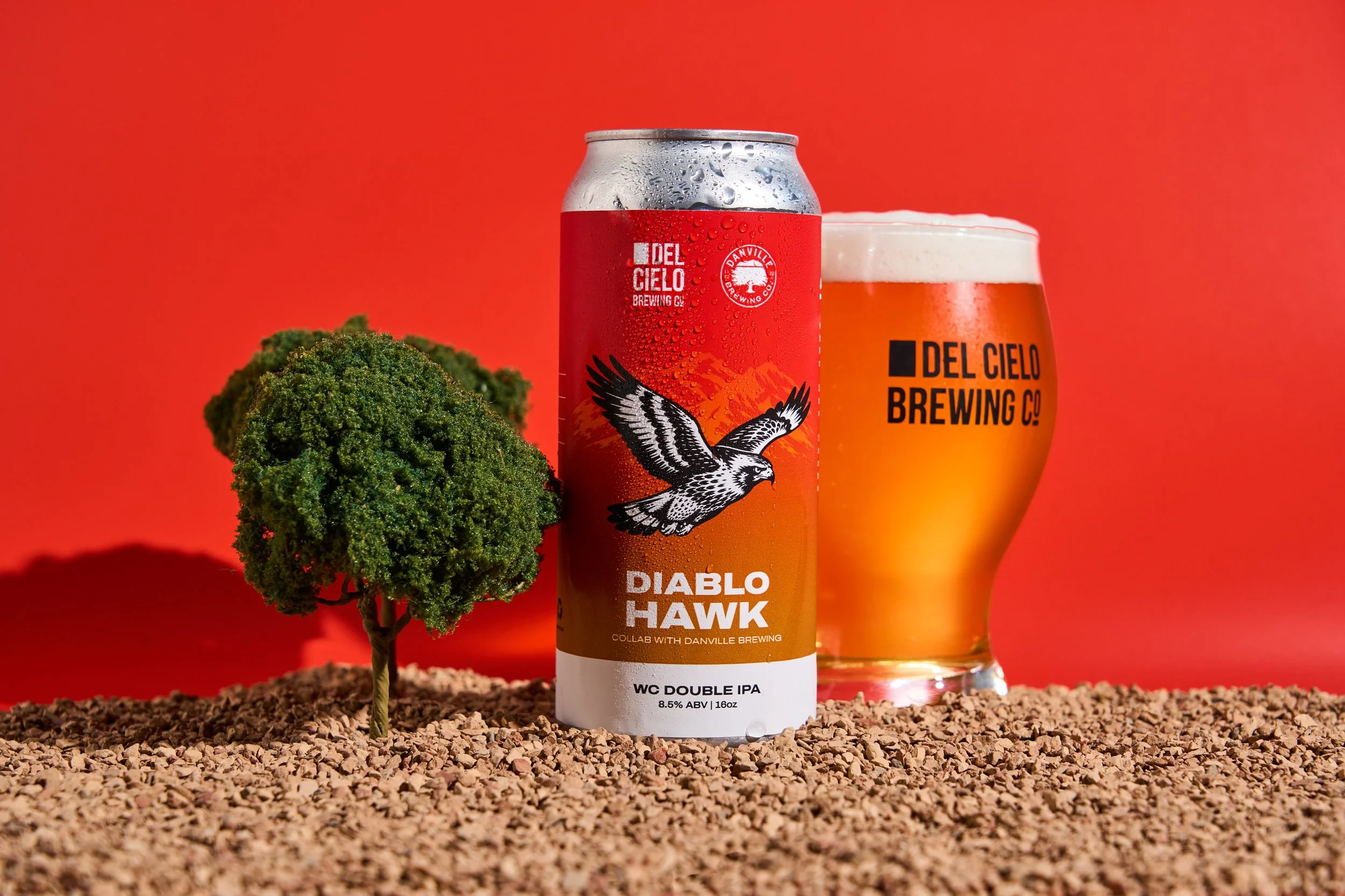 A can of Diablo Hawk beer from Del Cielo Brewing Co. alongside a glass of beer, with a small artificial tree and a dirt-like surface on a red background.