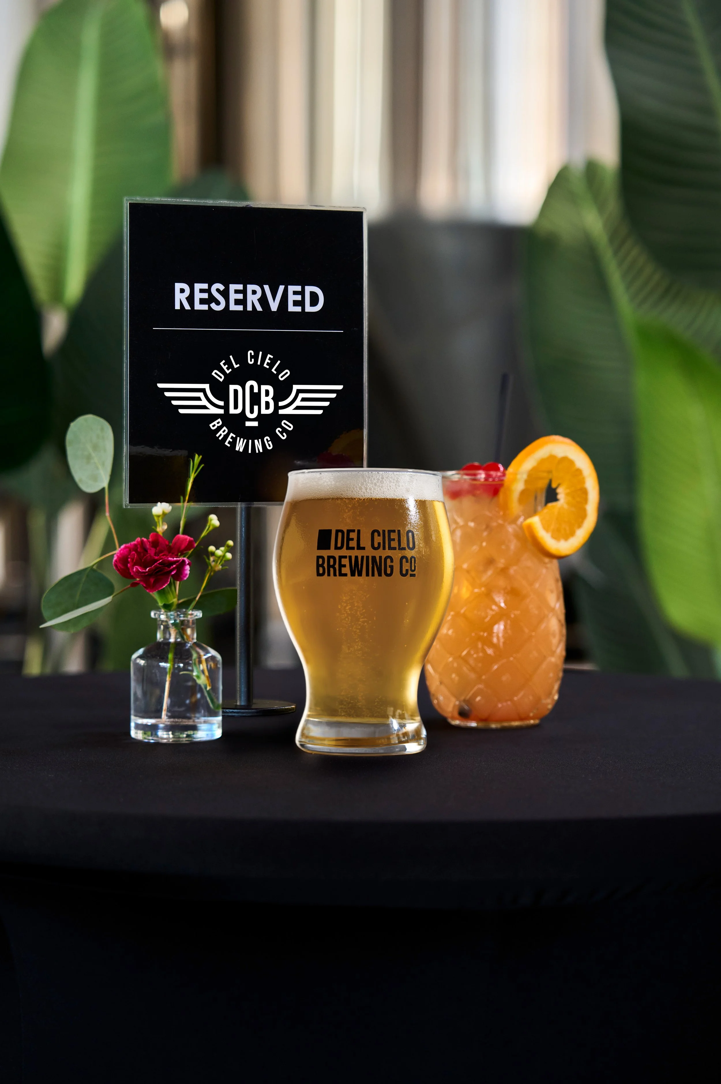 A table with two beers from Del Cielo Brewing Co, one in a tall glass and the other in a large glass with orange slices and red berries, alongside a small vase with pink and white flowers, a reserved sign for the brewery, and tropical green foliage in the background.
