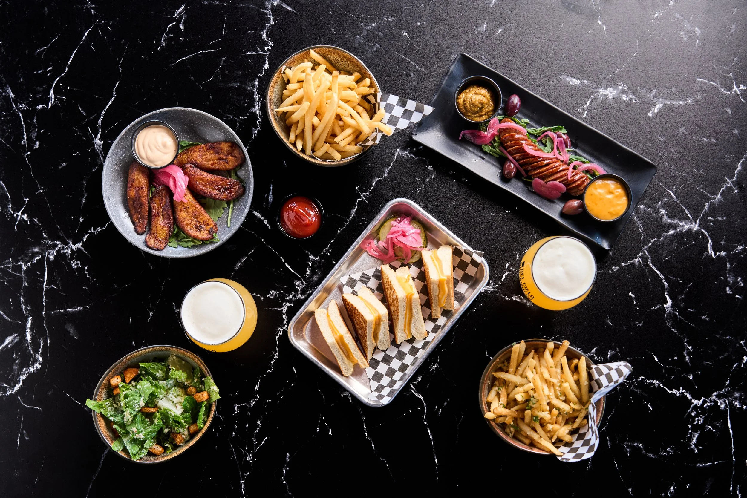 Various dishes including fries, plantains with dipping sauces, sliced sausage with pickled onions and greens, a salad, sandwiches, and glasses of beer on a black marble table.
