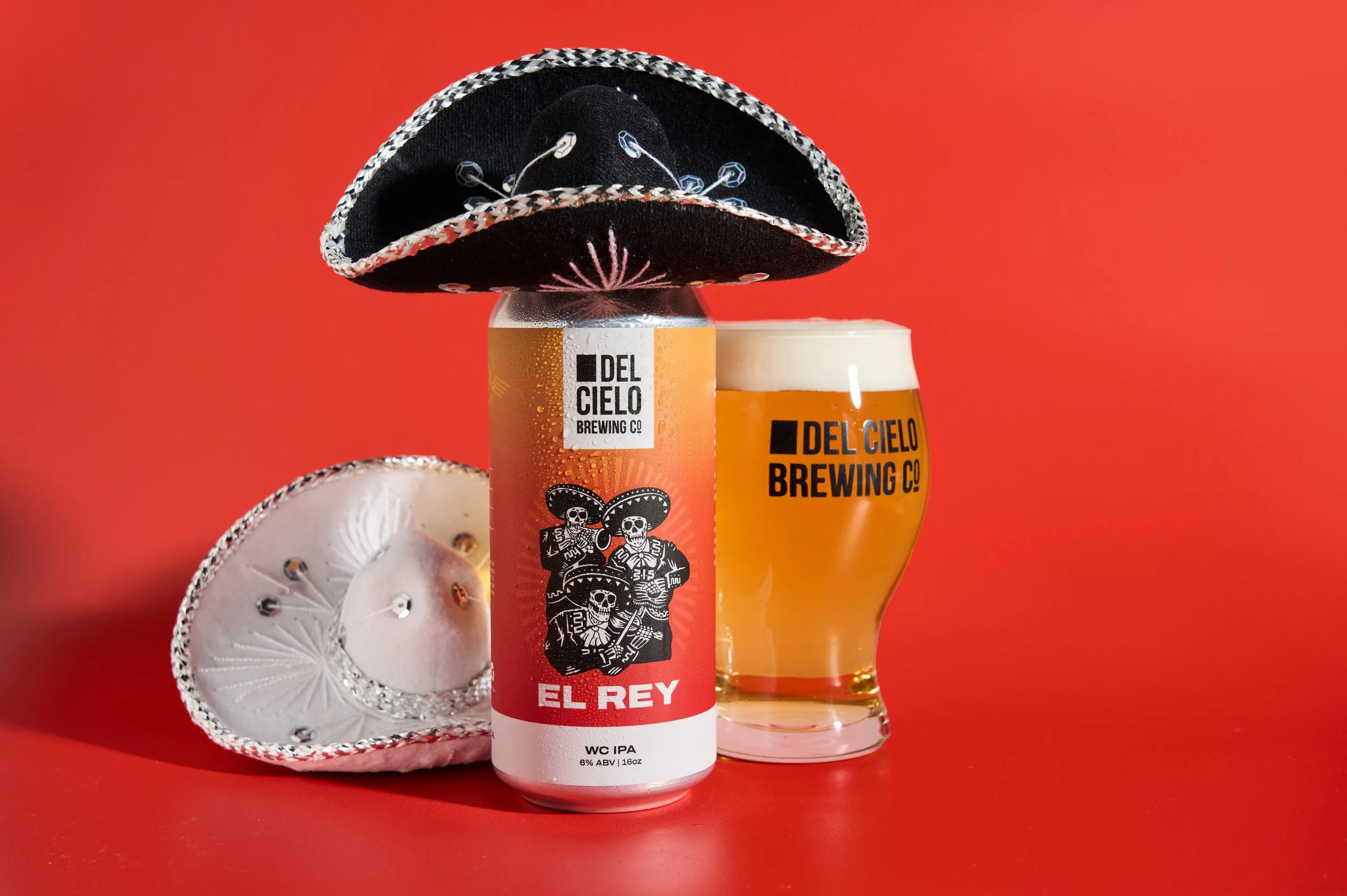 A can of El Rey beer with a sombrero placed on top, a glass of beer beside it, and embroidered white sombrero with silver accents on a red background.