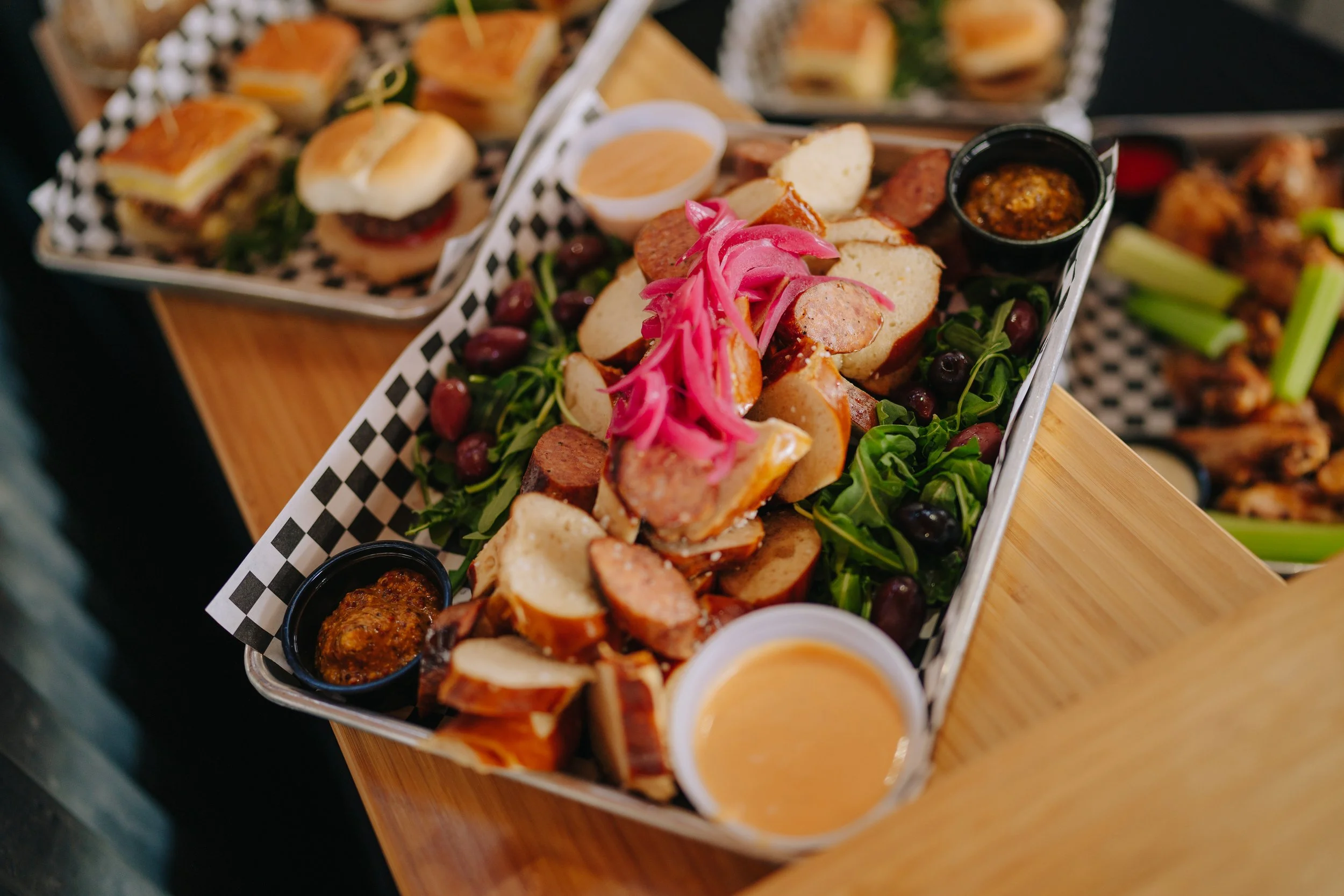 A tray of assorted appetizers including mini sandwiches, sausage and bread slices, pickled onions, olives, and dipping sauces in small containers.