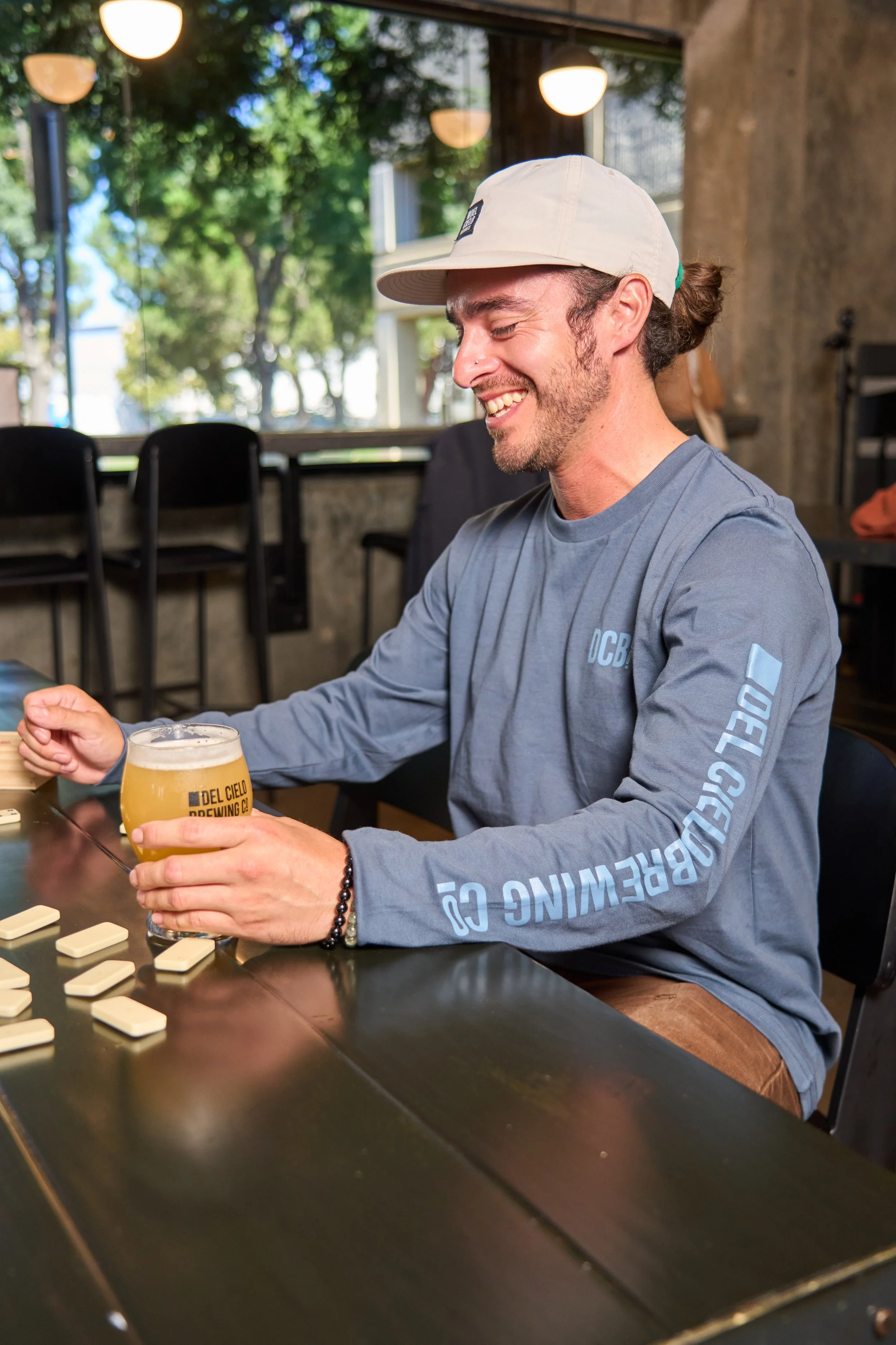 A man sitting at a table playing a game of dominoes, holding a beer glass, smiling, in a restaurant or cafe with large windows and trees outside.